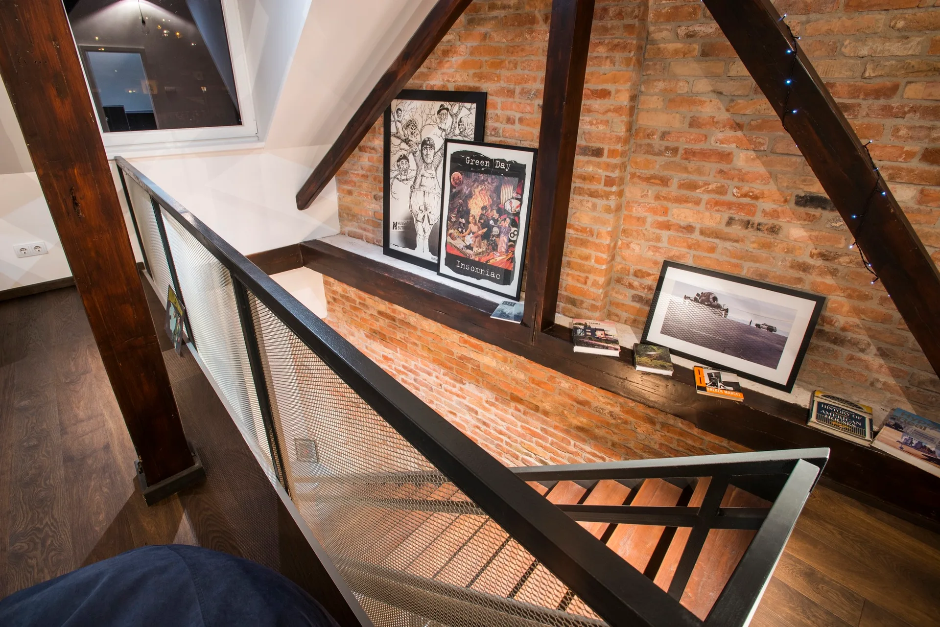A loft staircase landing with a black wire mesh railing, an exposed brick wall, and art pieces resting on a dark wood ledge.