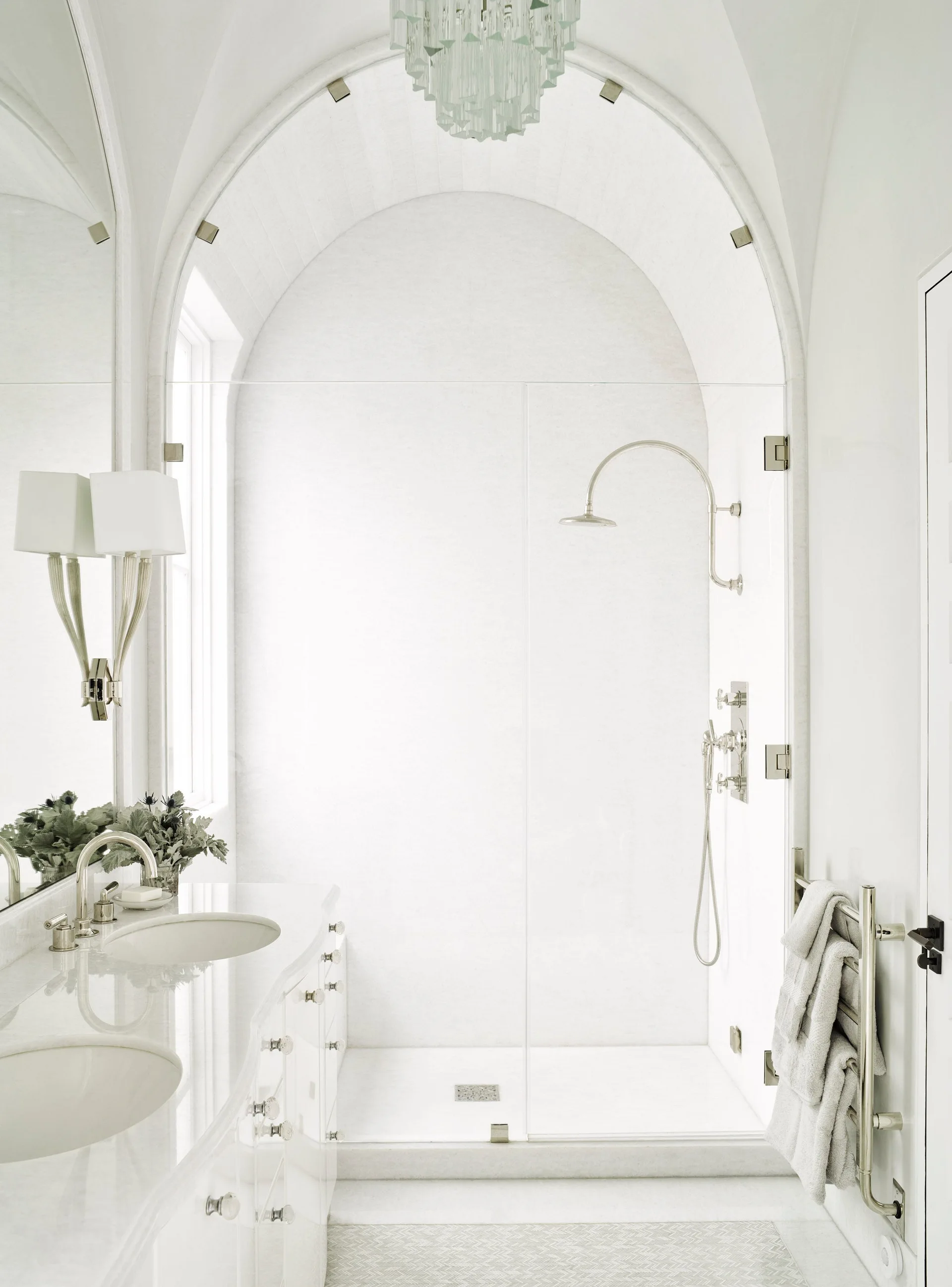 A bright white bathroom features a glass-enclosed walk-in shower under an arched ceiling next to a dual-sink vanity with polished fixtures.