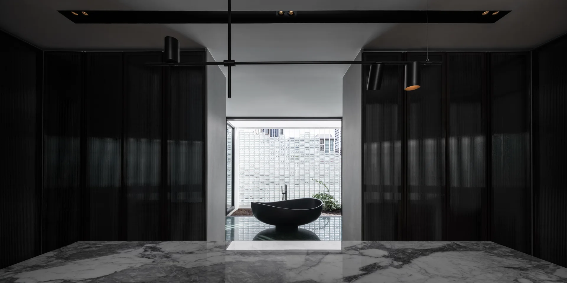 A modern dark bathroom with a black sculptural freestanding bathtub positioned in front of a bright glass block window and framed by dark fluted walls.