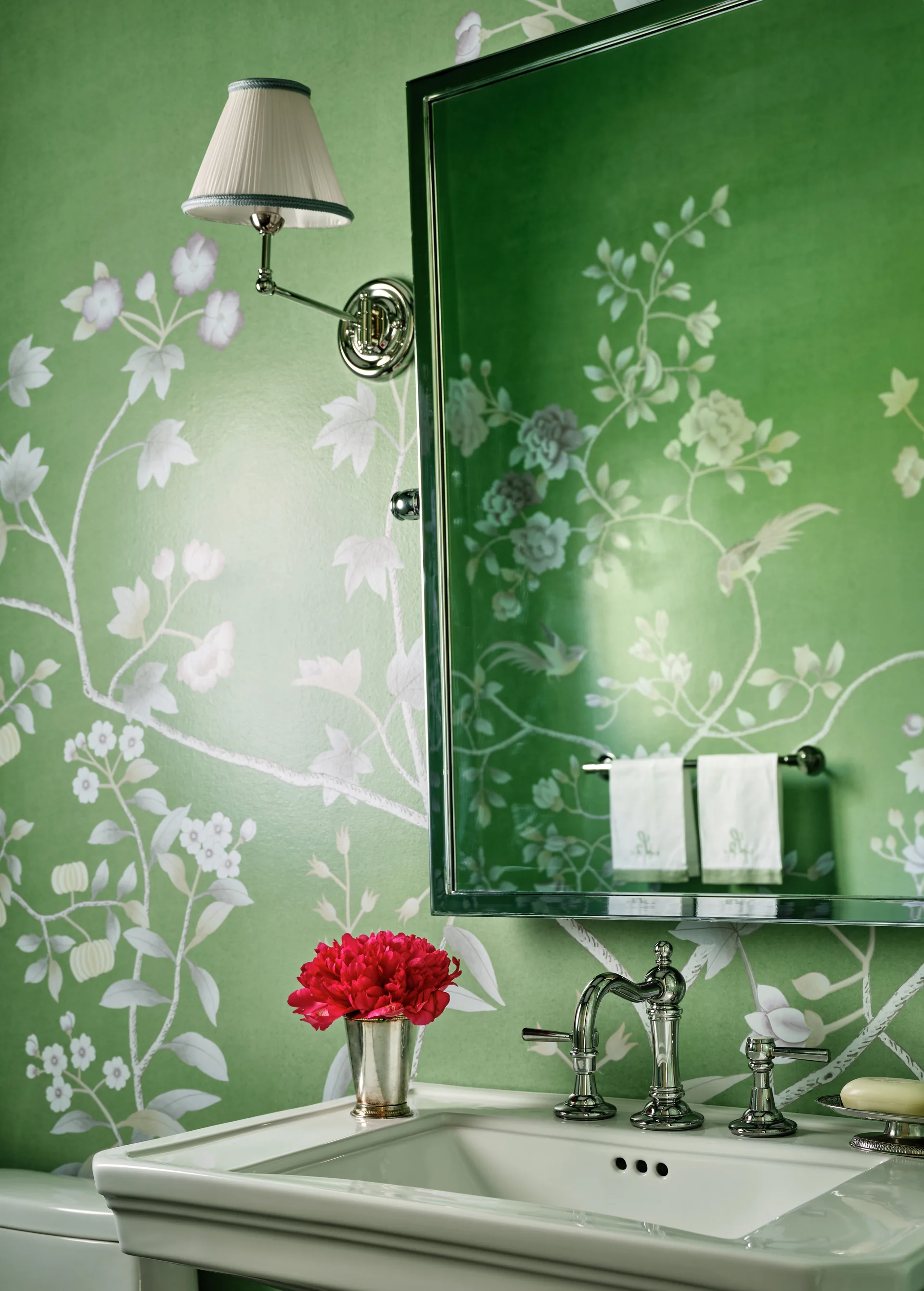 A close-up of a powder room vanity featuring a white sink, chrome fixtures, a matching wall sconce, and green botanical wallpaper.