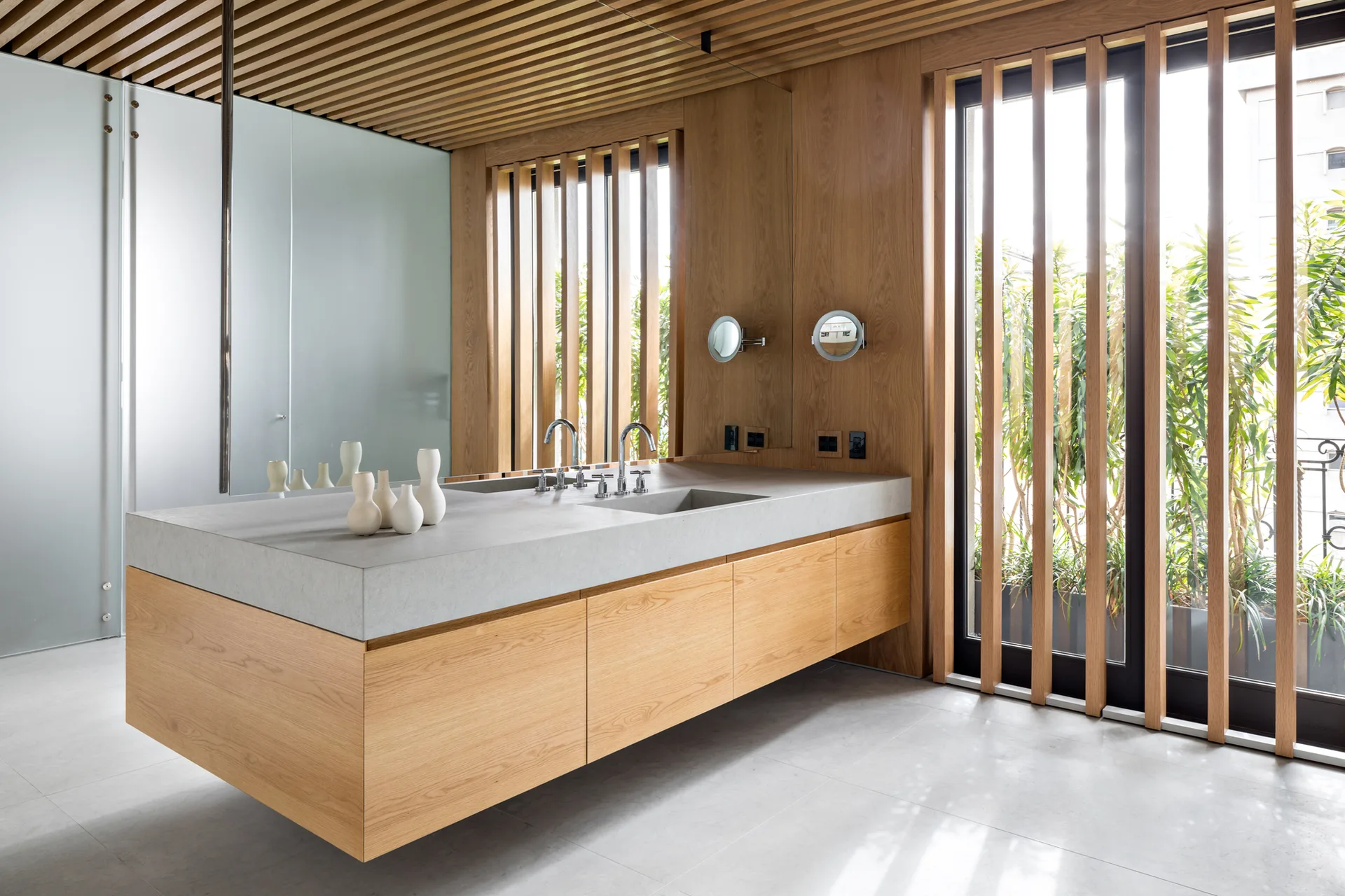 A minimalist bathroom featuring a floating light wood vanity with a thick grey concrete countertop, dual faucets, and vertical wood ceiling and window slats.