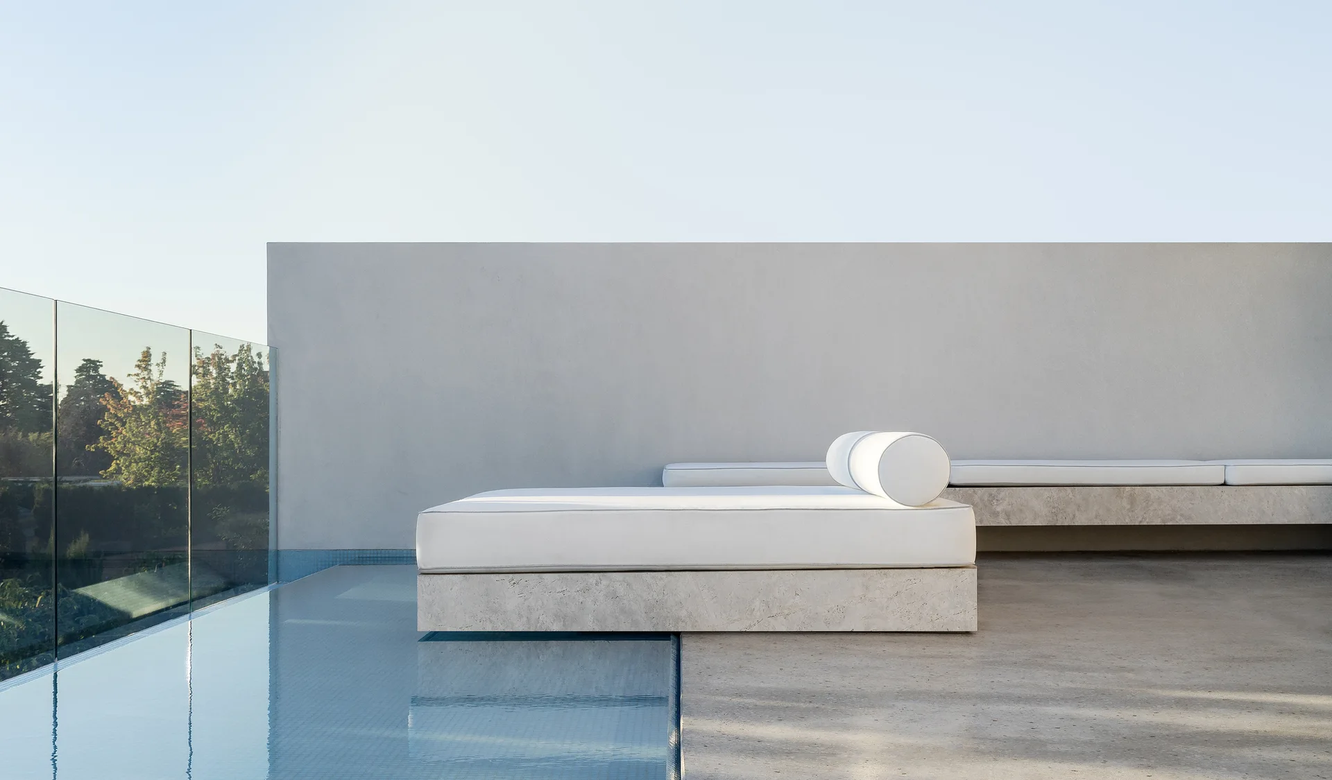 A minimalist outdoor daybed with a light stone base and white cushions sits beside a pool with a glass railing against a grey wall.