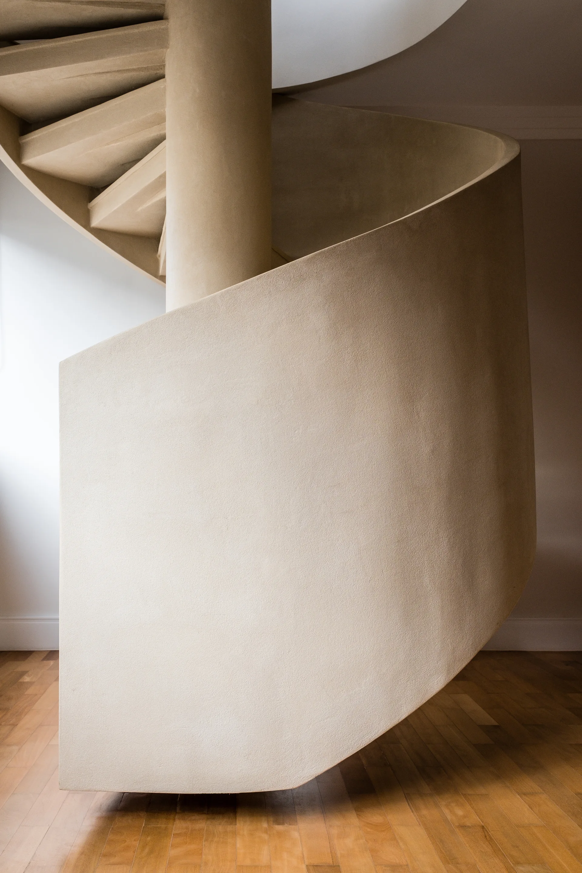 A sculptural beige spiral staircase with a solid matte plaster finish curves upward from a light brown wood plank floor.