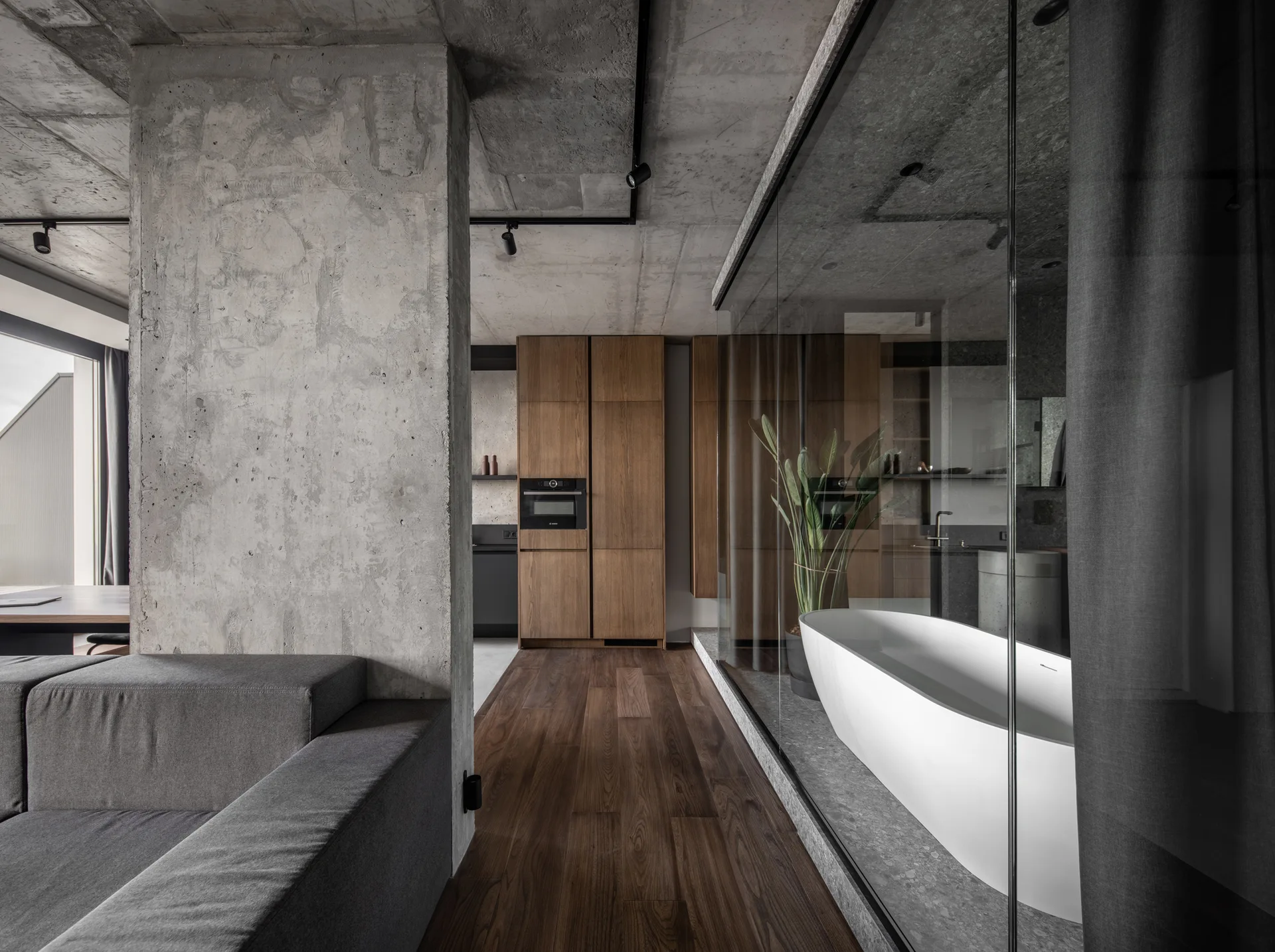 A modern loft interior features a raw concrete pillar, a freestanding white bathtub behind a glass partition, and warm wood flooring.