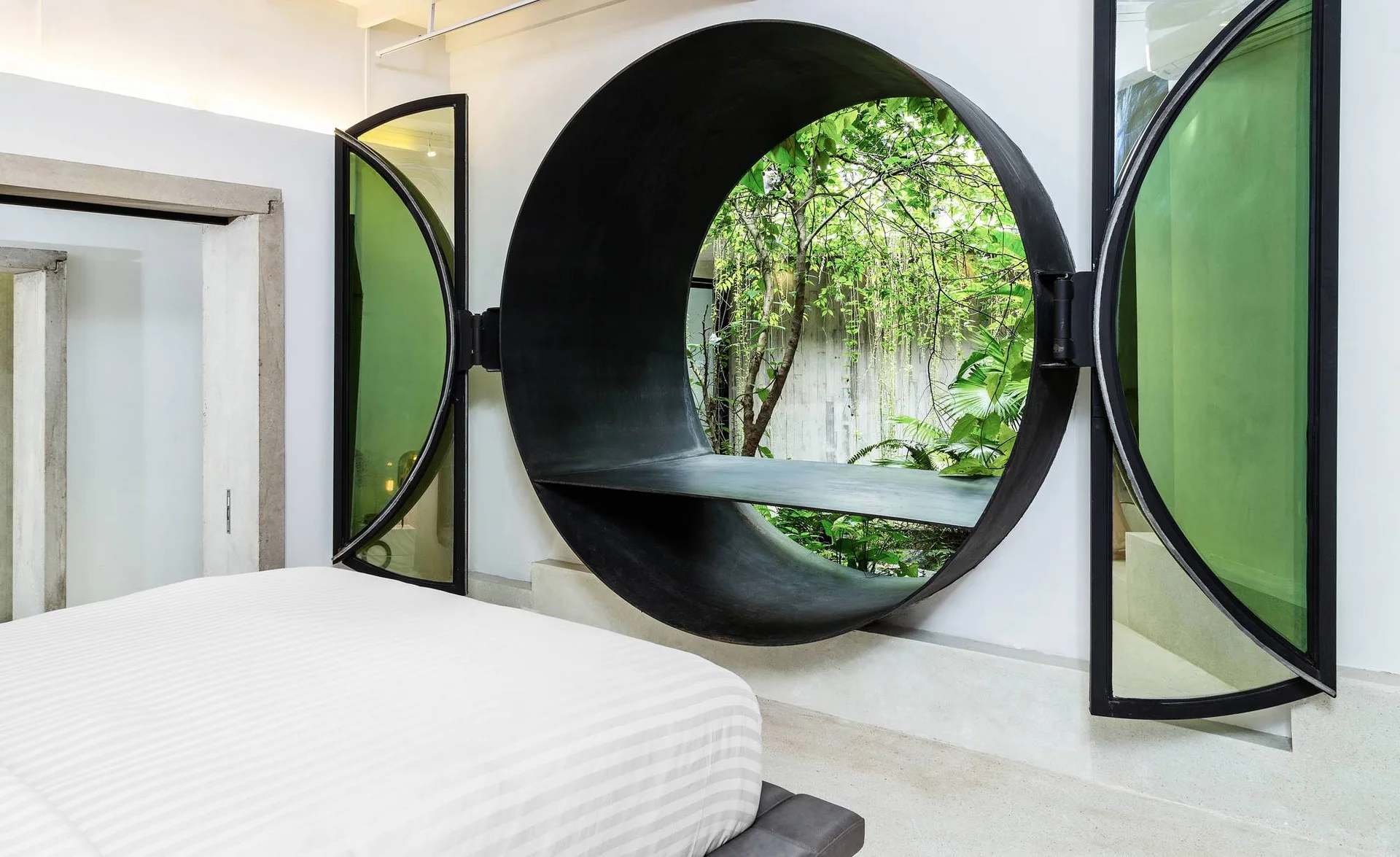 A modern bedroom with white striped bedding and a large circular metal window with hinged glass panels opening to an outdoor garden.