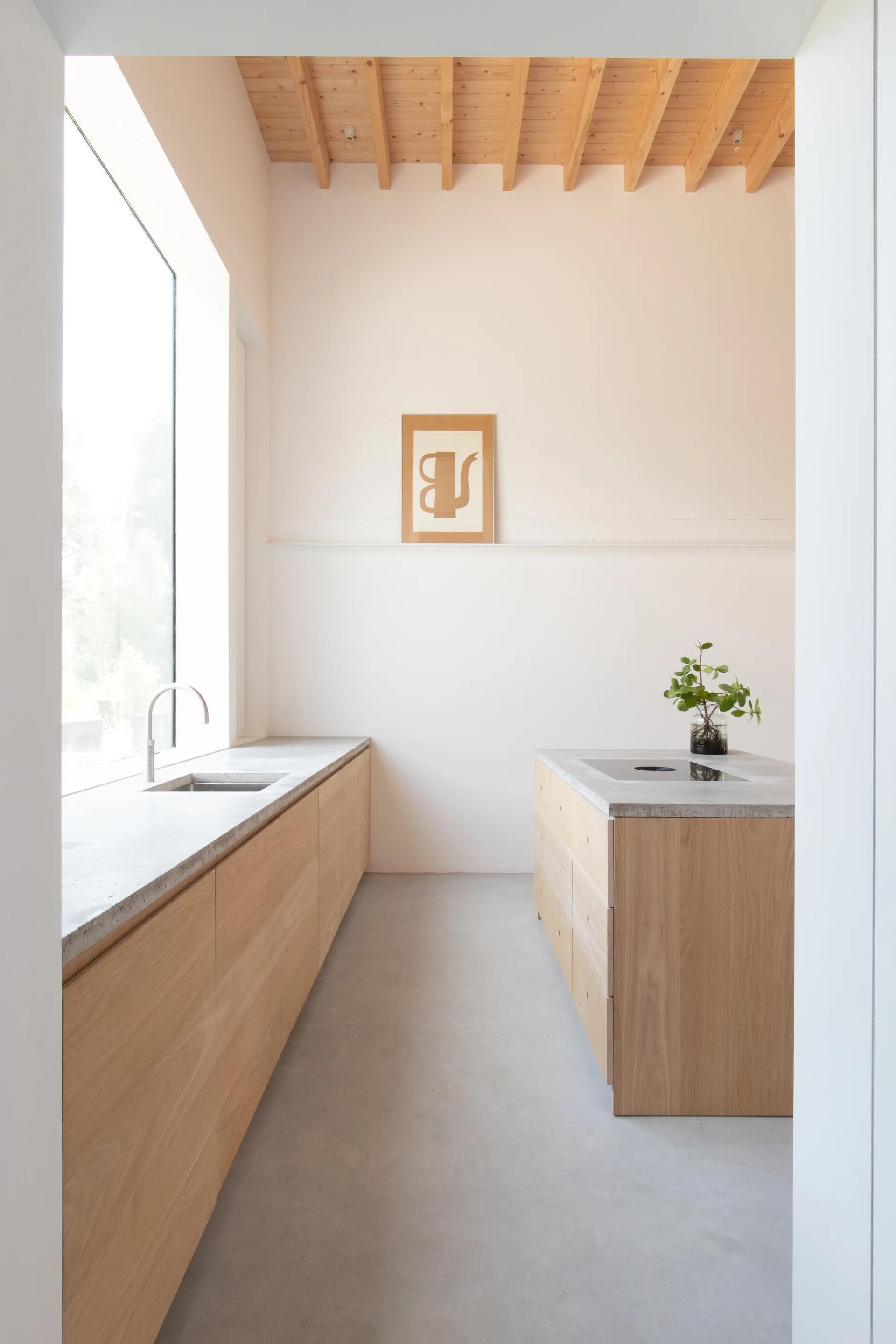 A minimalist kitchen features light wood cabinets, concrete countertops, a matching island, and an exposed timber beam ceiling.