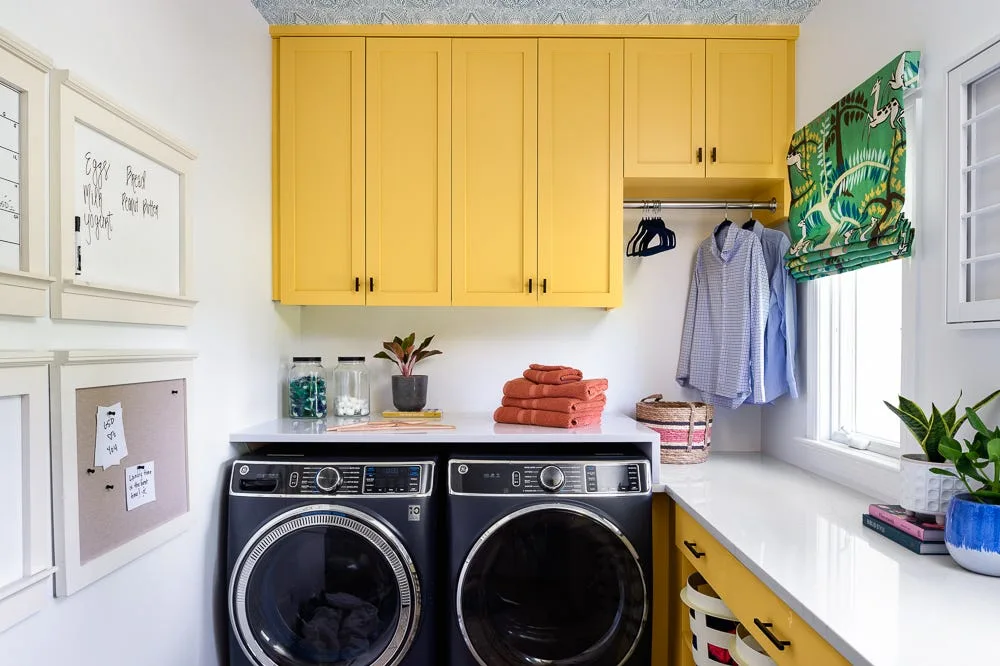 A modern laundry room with bright yellow cabinets, a white countertop over a black washer and dryer, and patterned wallpaper on the ceiling.