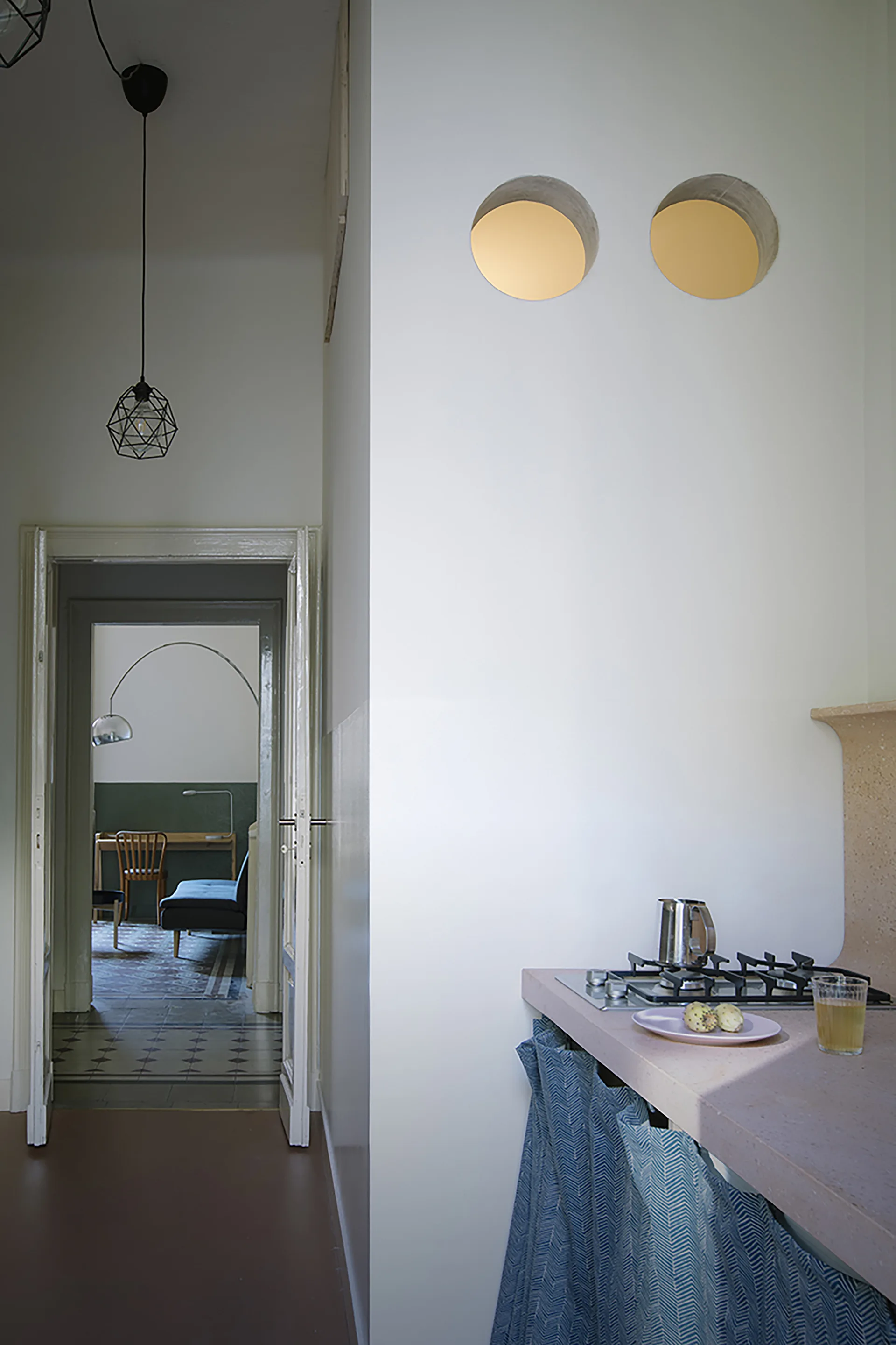 A kitchen with a pink stone counter, blue fabric skirt, and two circular wall openings looking through a doorway into a living area.