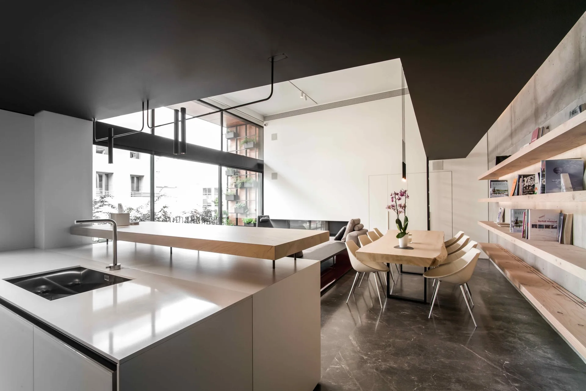 A modern kitchen and dining space features a white island with a suspended wood bar, a live-edge table, and floating shelves against a concrete wall.