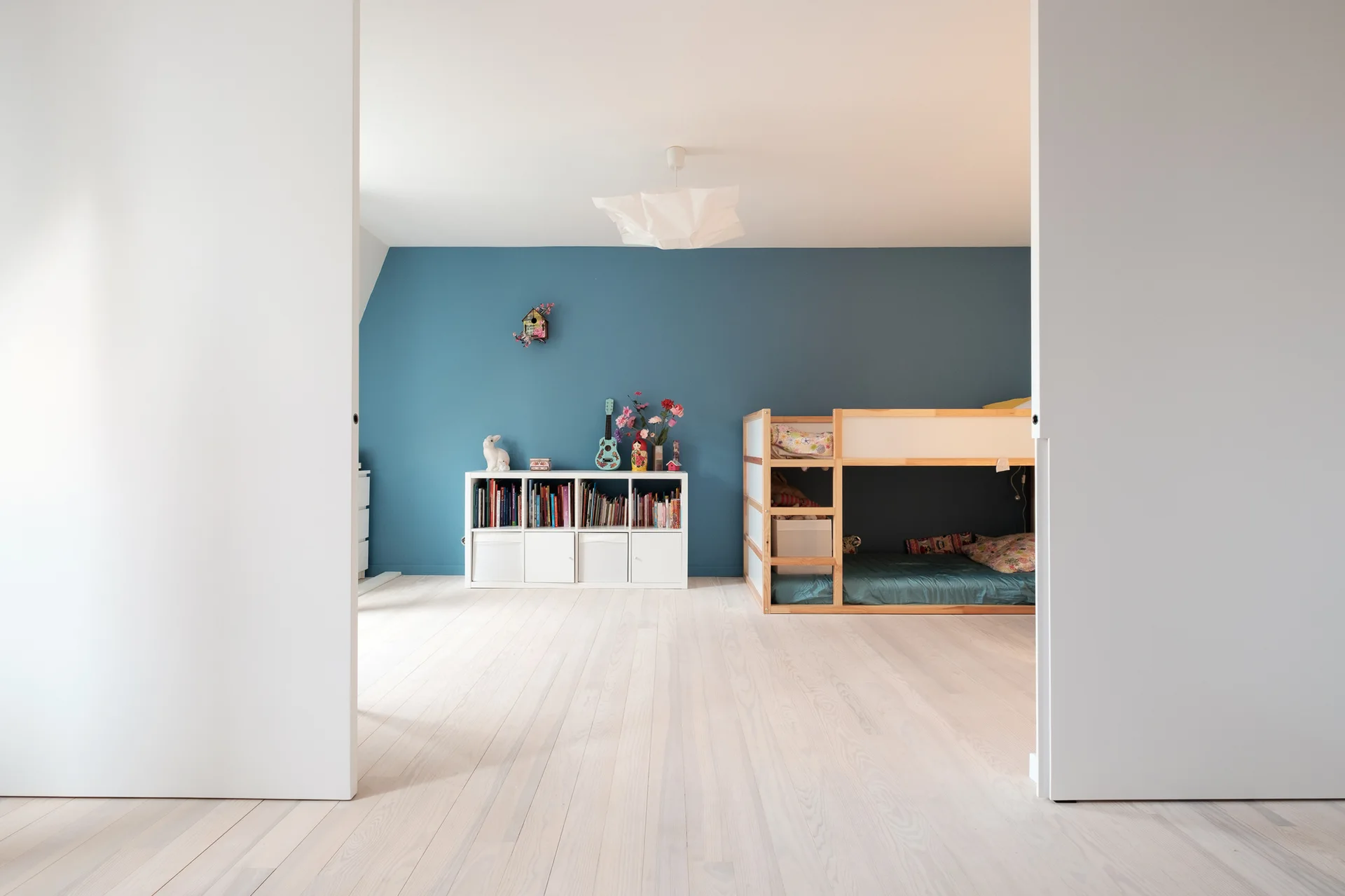 A child's bedroom viewed through open doors, featuring a blue accent wall, a light wood bunk bed, and a low white storage unit on pale wooden floors.