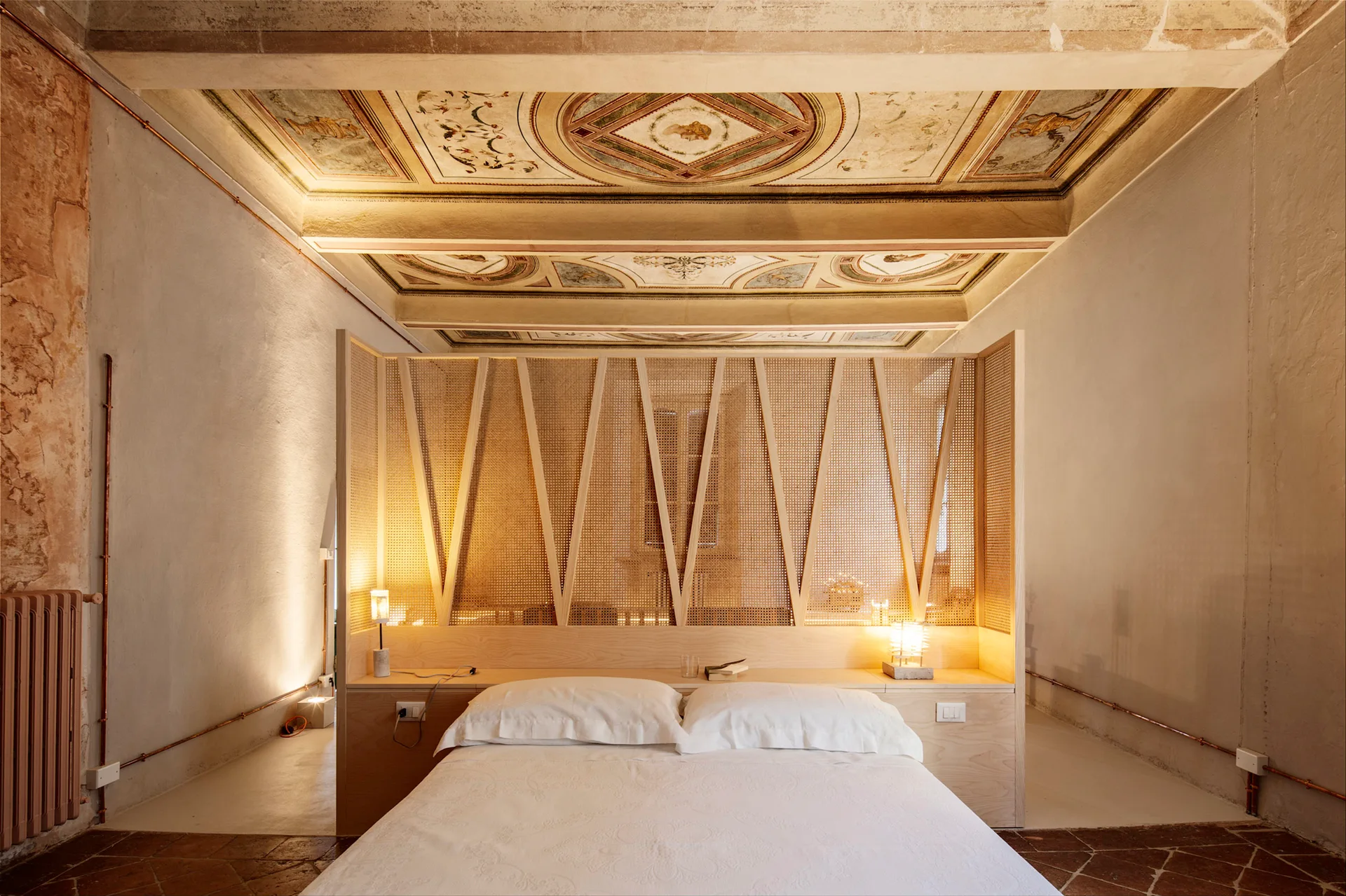 A low bed sits before a tall wood and cane partition wall in a historic bedroom featuring a highly detailed frescoed ceiling.