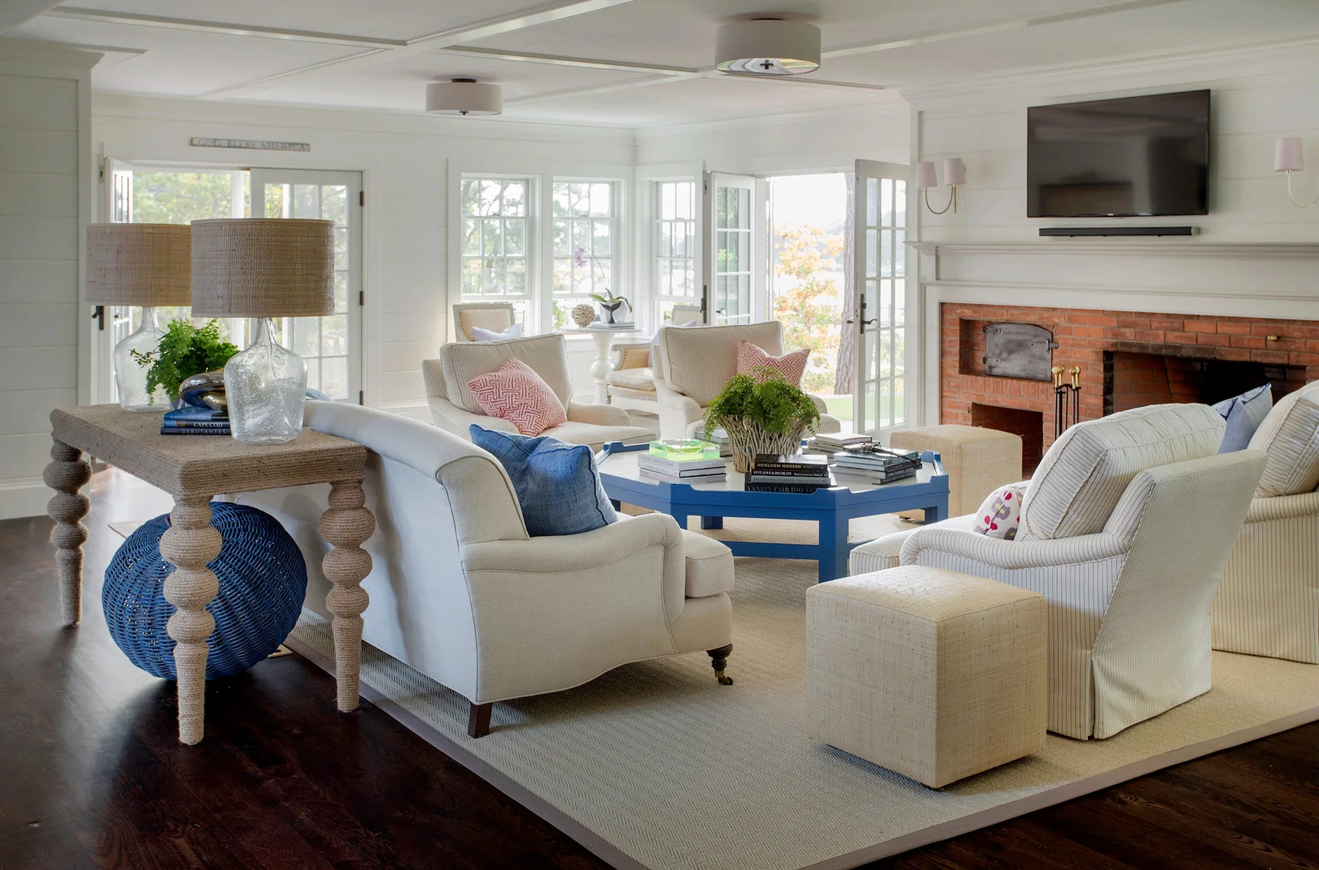 A bright coastal living room featuring a blue coffee table, white shiplap walls, two cream-colored sofas, and a brick fireplace.