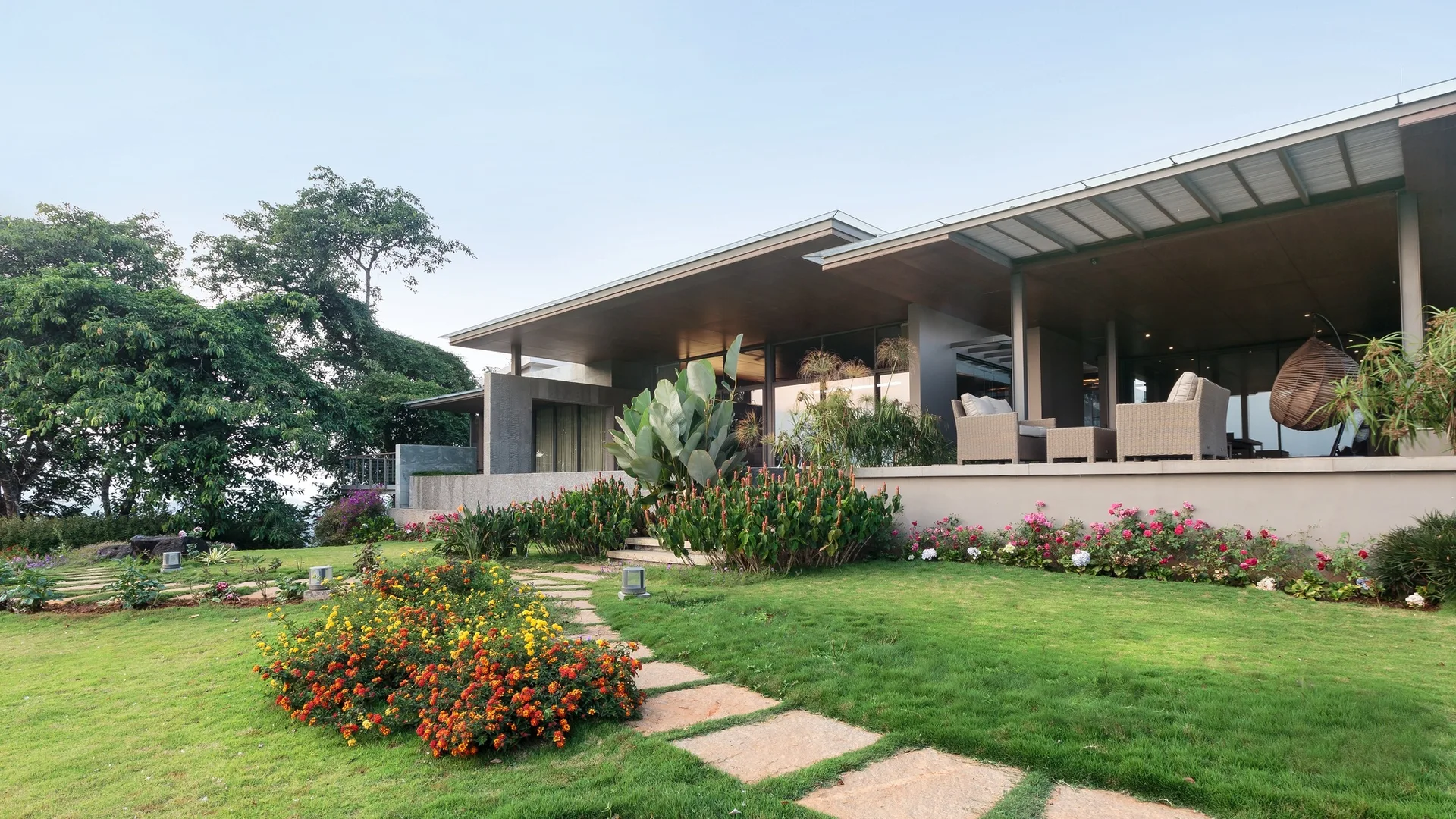 A modern covered terrace features wicker armchairs, a hanging egg chair, and a stepping stone path winding through a manicured green lawn with flowers.