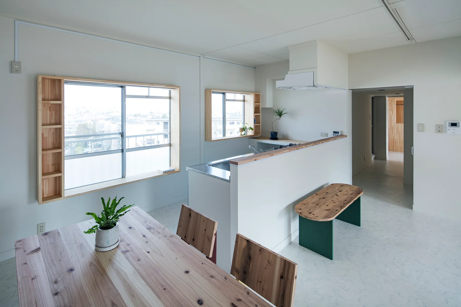 A minimalist white kitchen and dining area features light wood tables, a breakfast bar, and custom wooden shelving built around the window frames.
