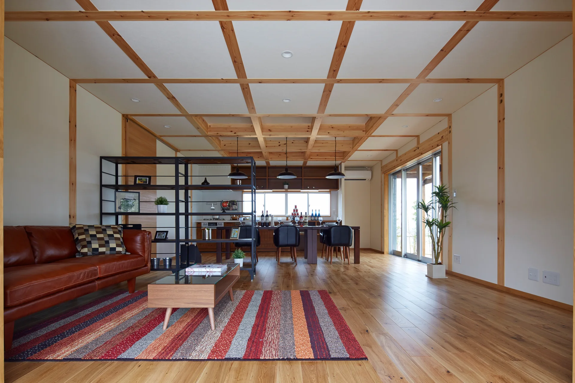 A modern open-concept living room features a brown leather sofa, striped rug, metal open shelving, and a grid pattern timber ceiling.