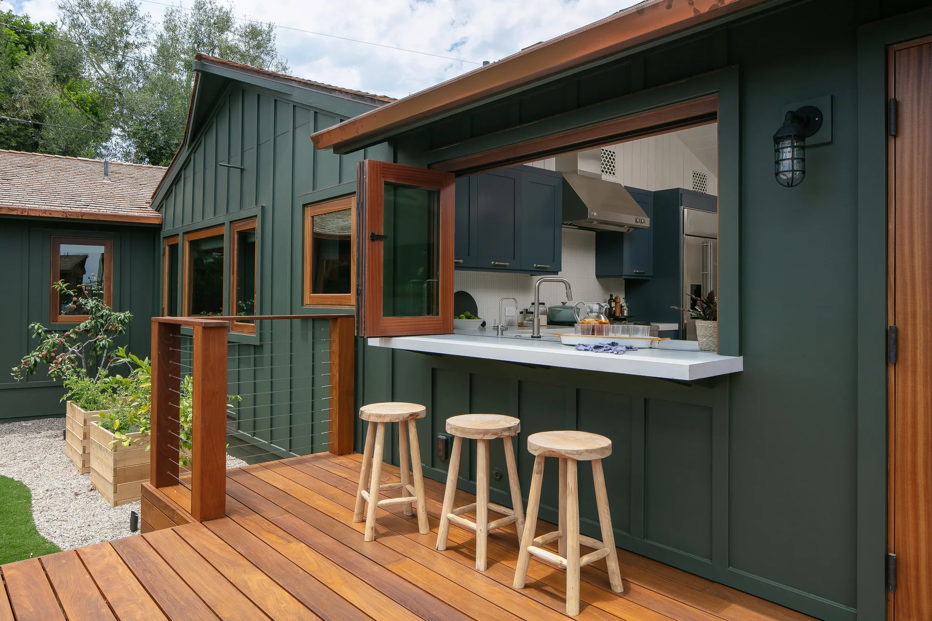 Three wooden bar stools sit on a wooden deck next to a pass-through window with a white counter leading into an indoor kitchen.