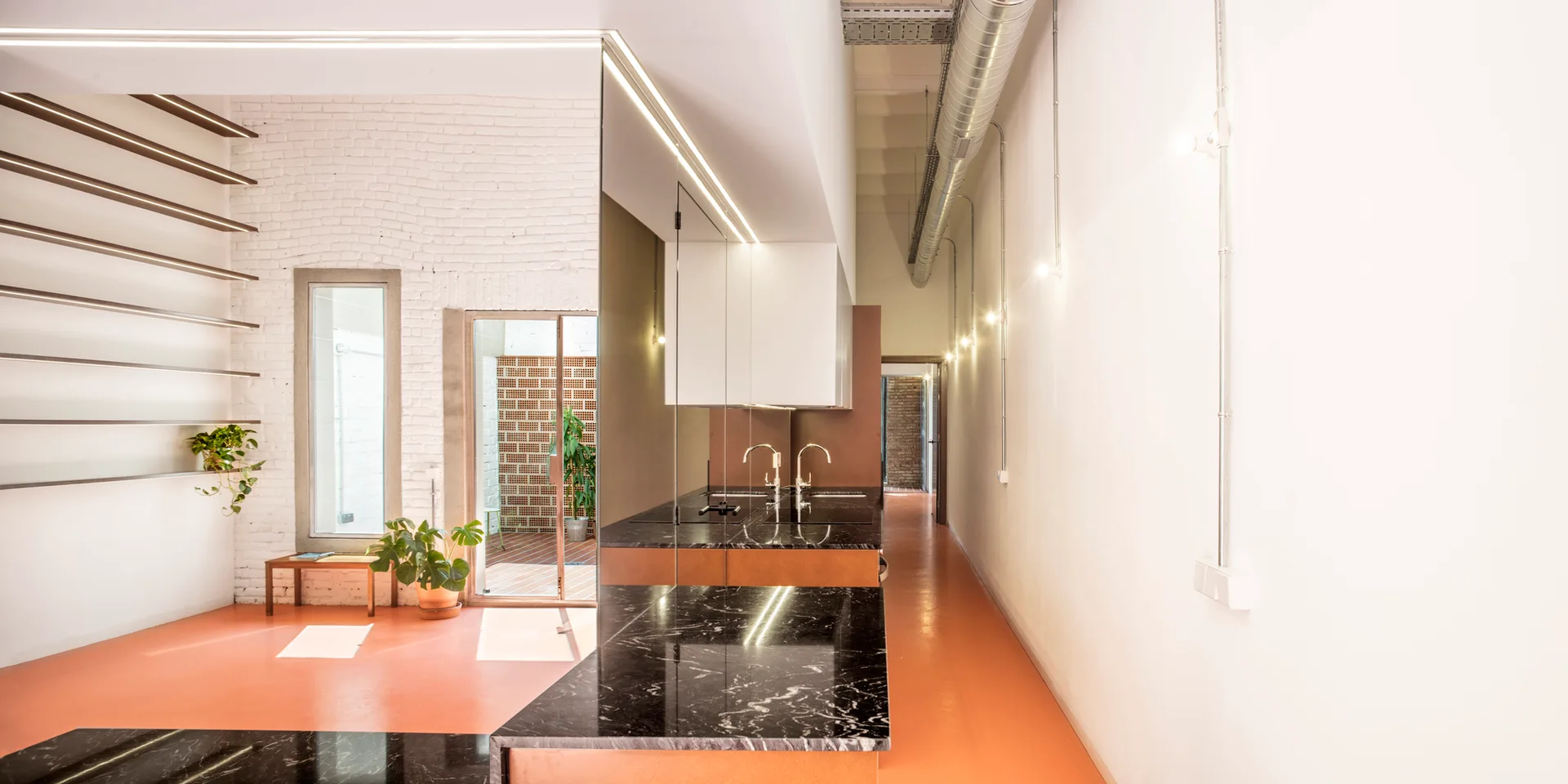 A modern industrial kitchen features a black marble island, a terracotta floor, white brick walls, and exposed overhead metal ductwork.