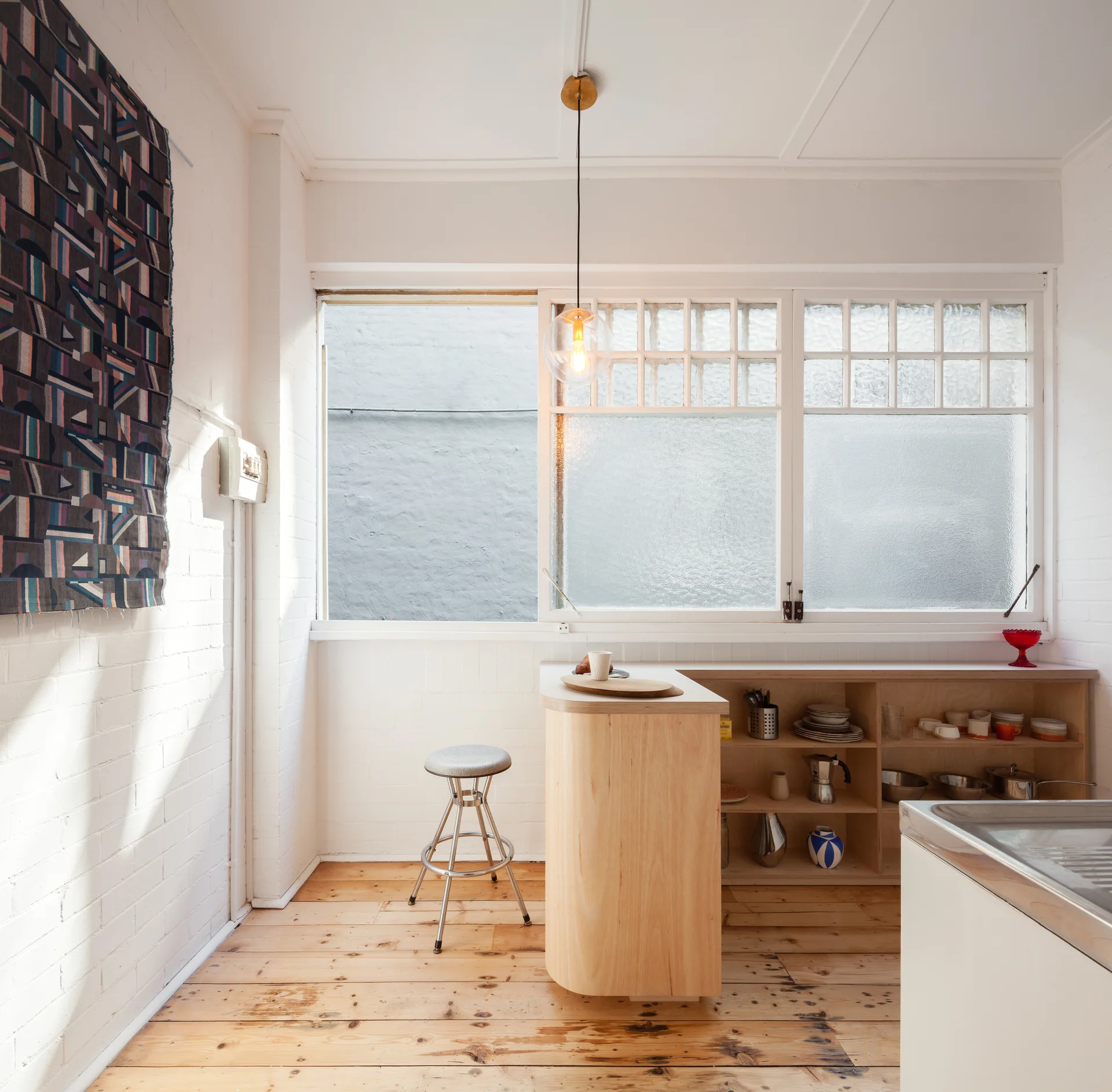 A minimalist kitchen features a curved light wood island, open shelves, a metal stool, and large frosted glass windows against painted brick walls.