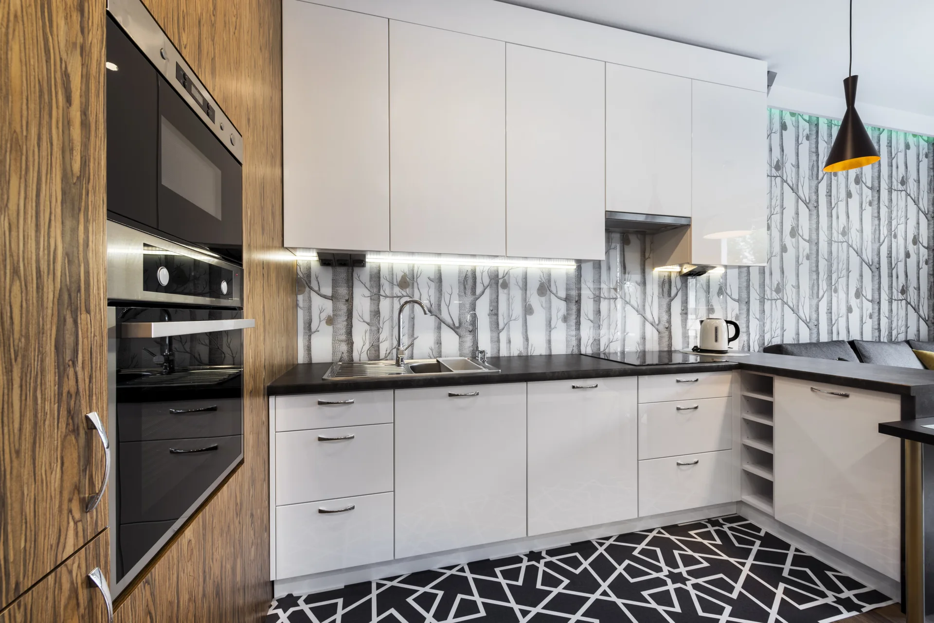 A modern kitchen featuring glossy white cabinets, a dark countertop, a forest-themed backsplash, and geometric black-and-white flooring.