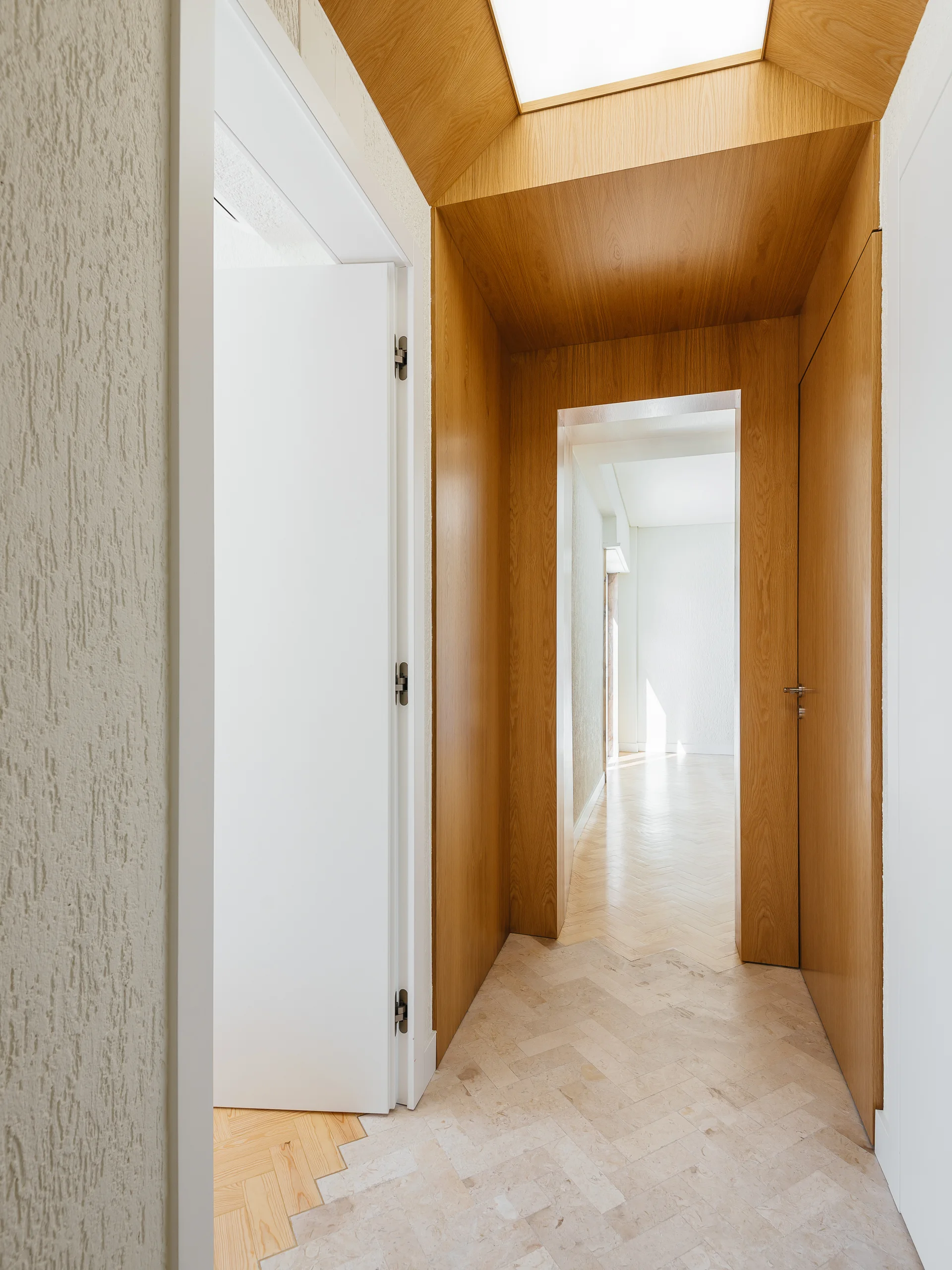 A modern residential hallway featuring a wood-wrapped portal, herringbone tiled flooring, and a white open door leading to an adjacent room.