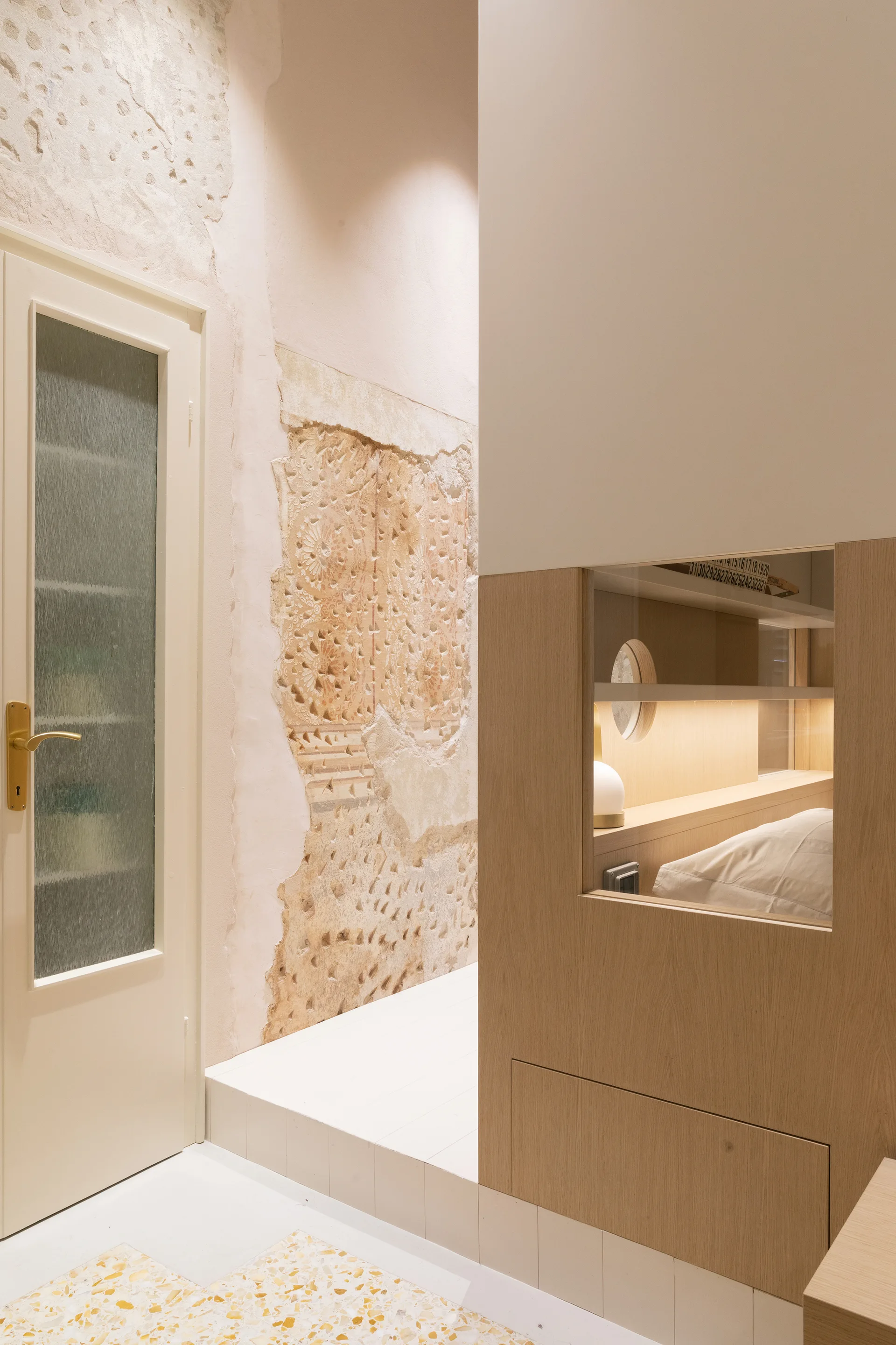 A light wood built-in bedroom enclosure stands next to an exposed plaster relief wall and a cream door with a glass panel.