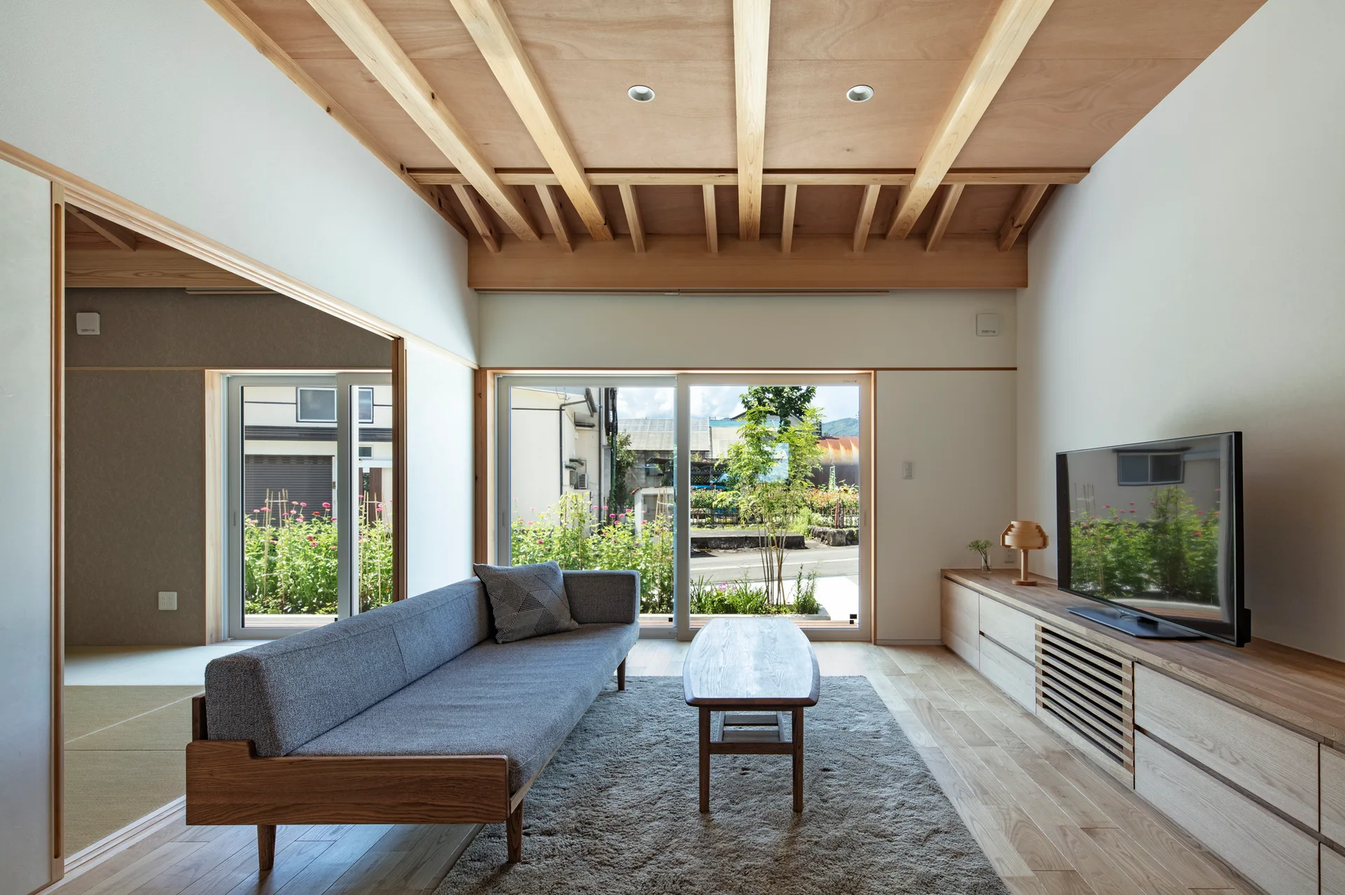 A minimalist living room features a grey sofa, wooden coffee table, and an exposed timber beam ceiling looking out to a green garden.