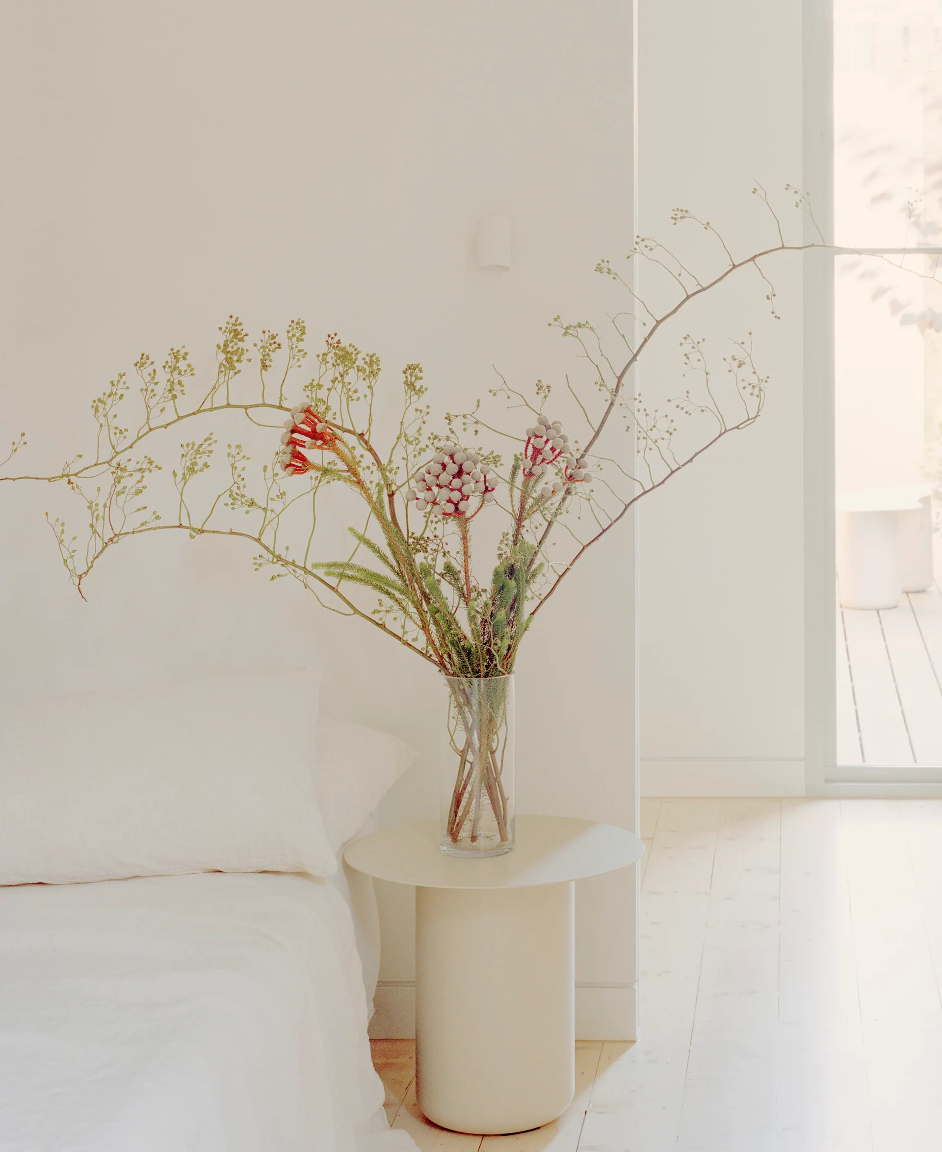 A white minimalist bedroom features a beige cylindrical side table topped with a glass vase of wild branches next to a bed with white linen bedding.