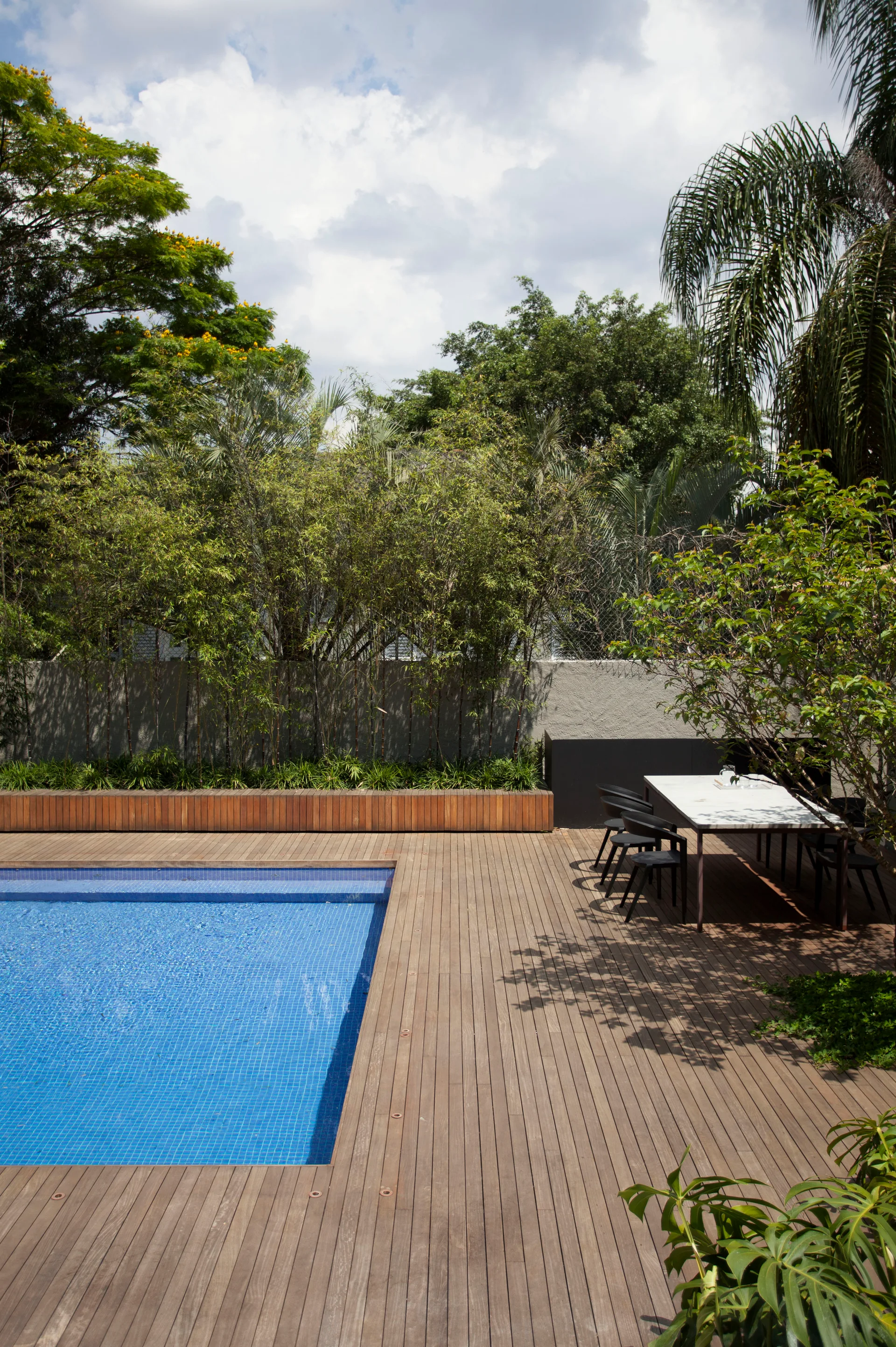 A blue tiled swimming pool is set within a wide wood plank deck next to an outdoor dining table and a raised planter with bamboo.