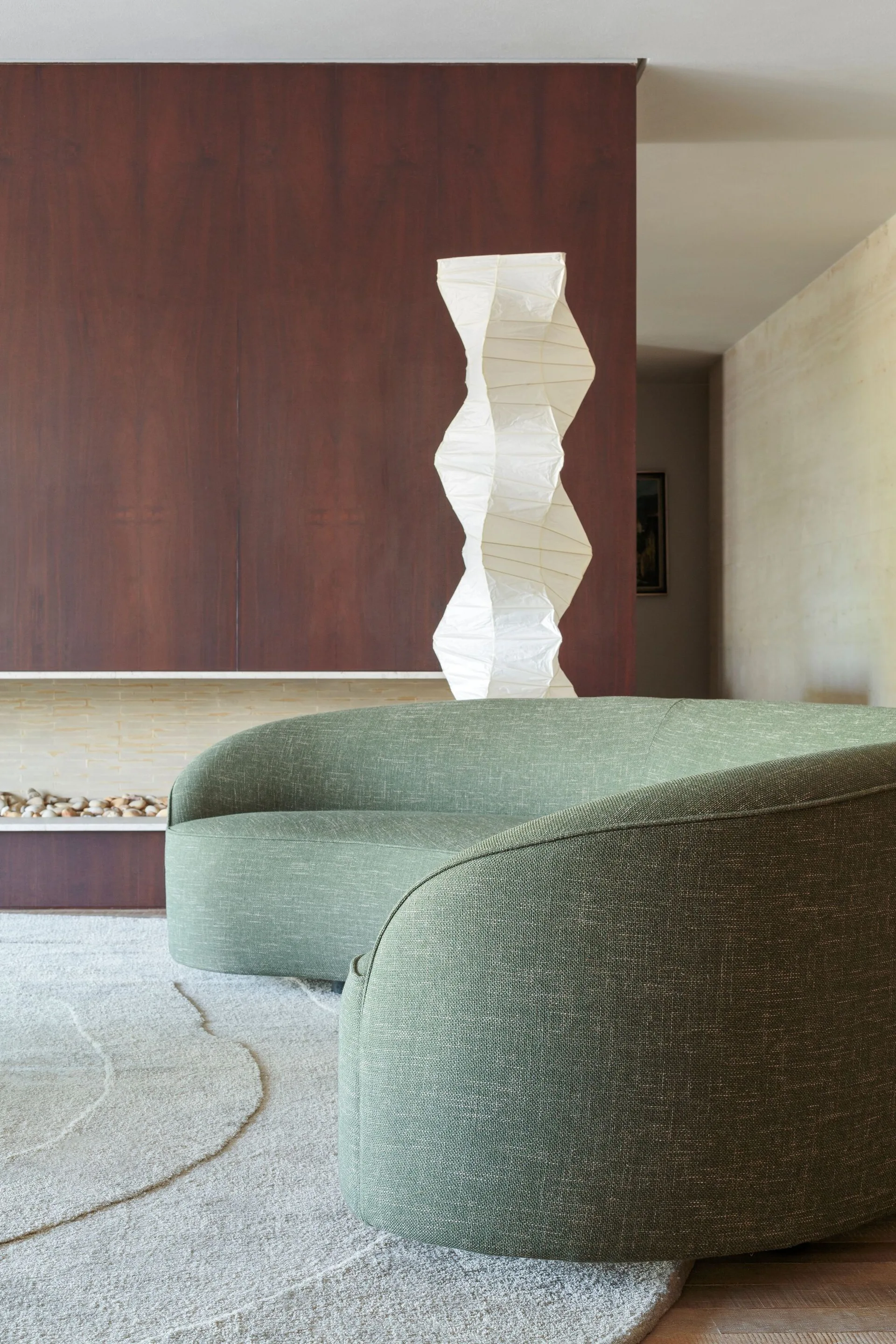 A curved green fabric sofa sits on a textured beige rug in front of a dark wood paneled wall and a sculptural white paper floor lamp.