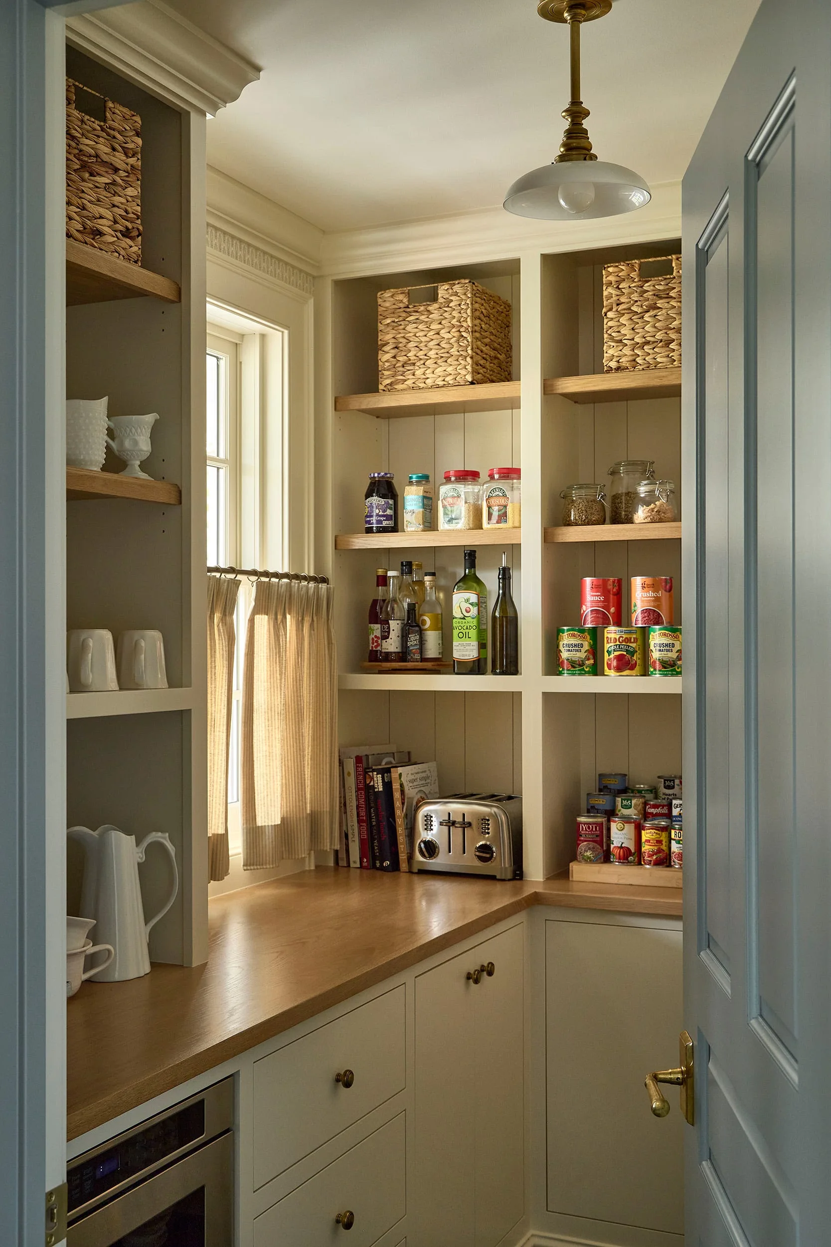 An organized cream kitchen pantry features built-in cabinets, warm wood countertops, open shelving with baskets and canned goods, and a small window.