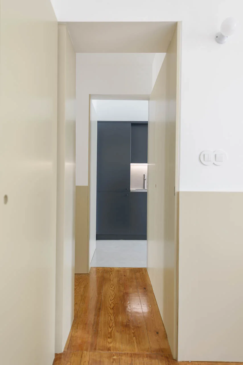 A narrow hallway with two-tone beige and white walls and wood flooring leads into a kitchen with dark charcoal cabinets.