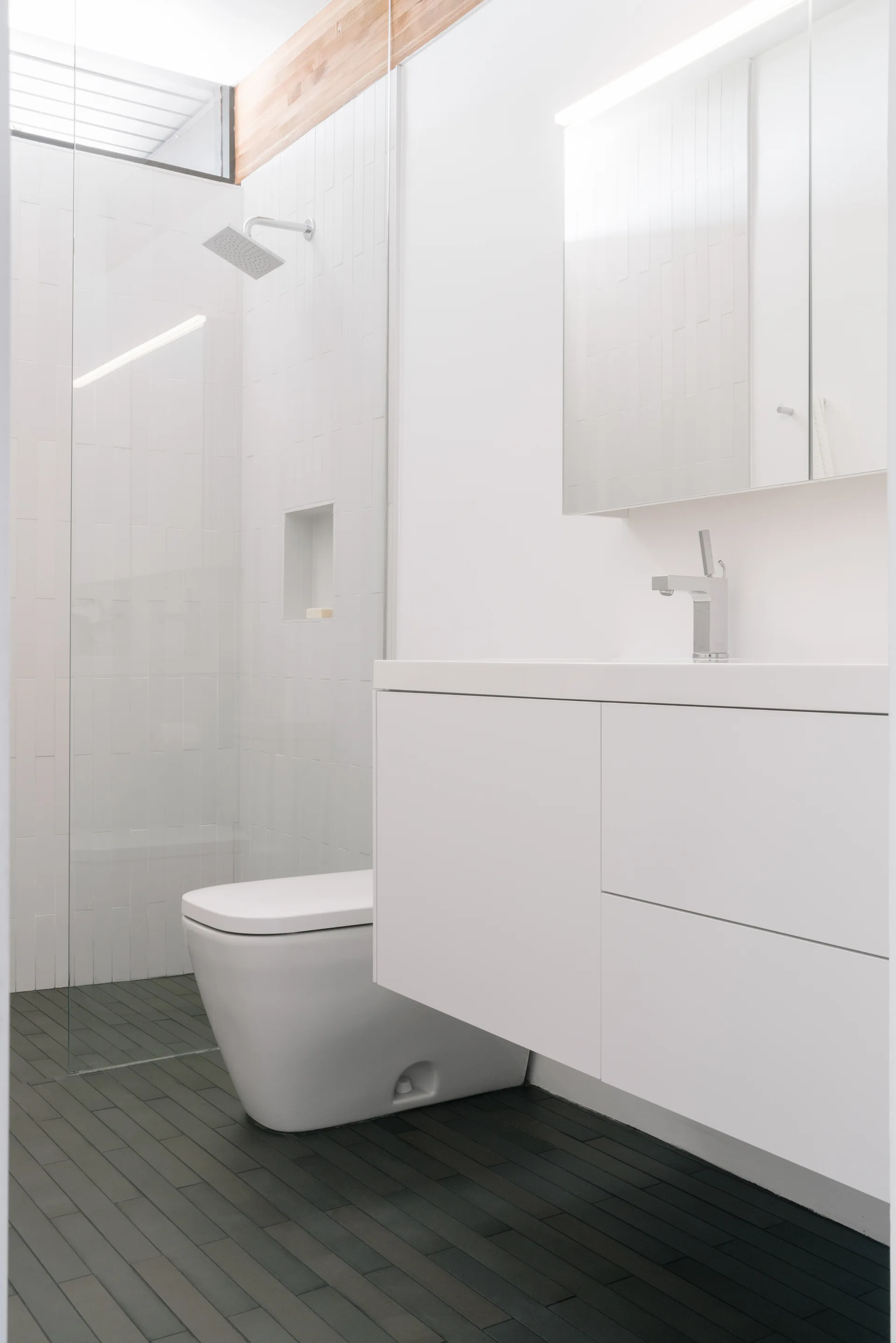 A minimalist white bathroom with a floating vanity, a glass shower partition, vertical wall tiles, and dark grey floor tiles.