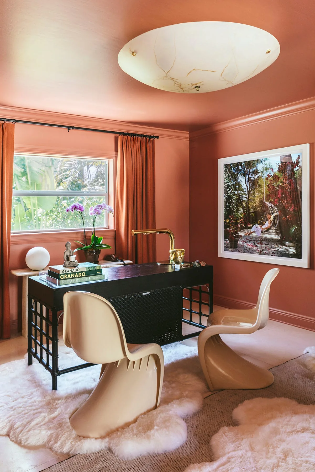 A home office features solid terracotta walls and ceiling, a central black desk with two cream S-chairs, and a large framed photograph on the wall.