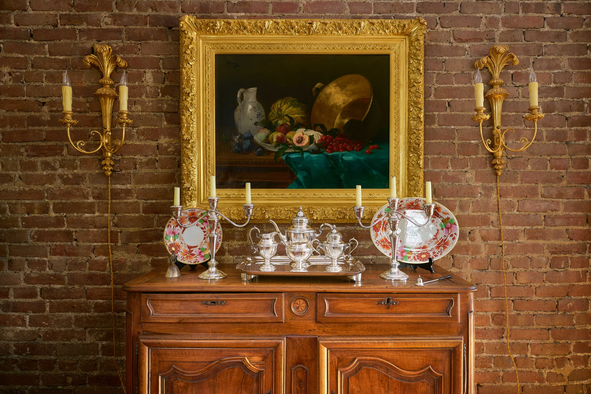 An antique wooden cabinet features a silver tea service and candelabras beneath a gold-framed painting on an exposed brick wall.