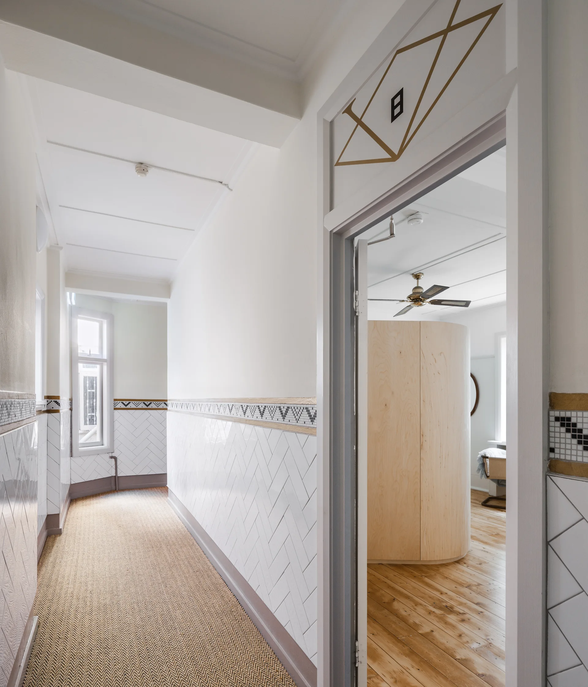 A white hallway featuring white herringbone tiled wainscoting and a woven runner leads past a doorway showing a curved plywood partition inside.