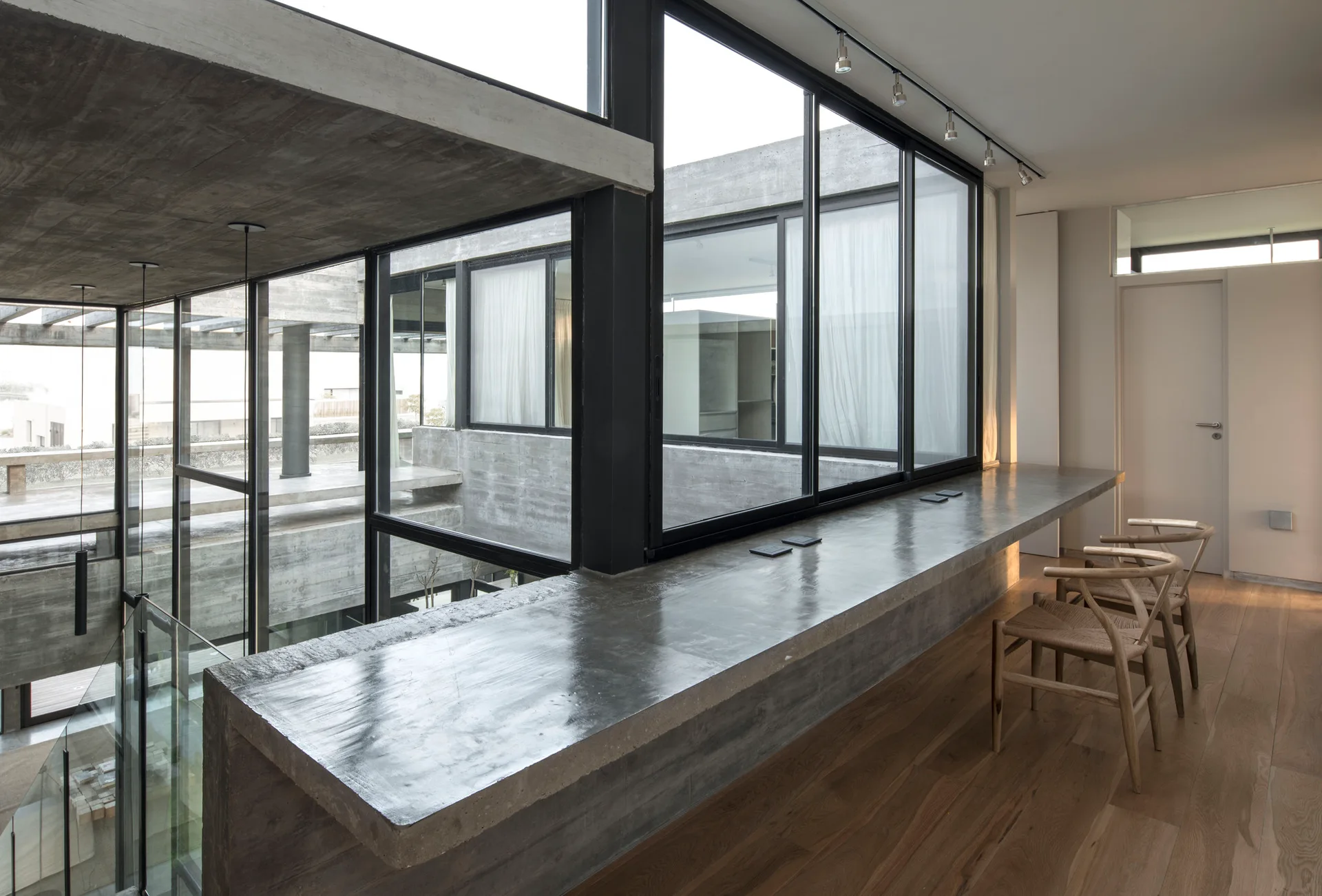 A modern industrial hallway with a long polished concrete desk built alongside large windows, featuring wooden chairs and an exposed concrete ceiling.