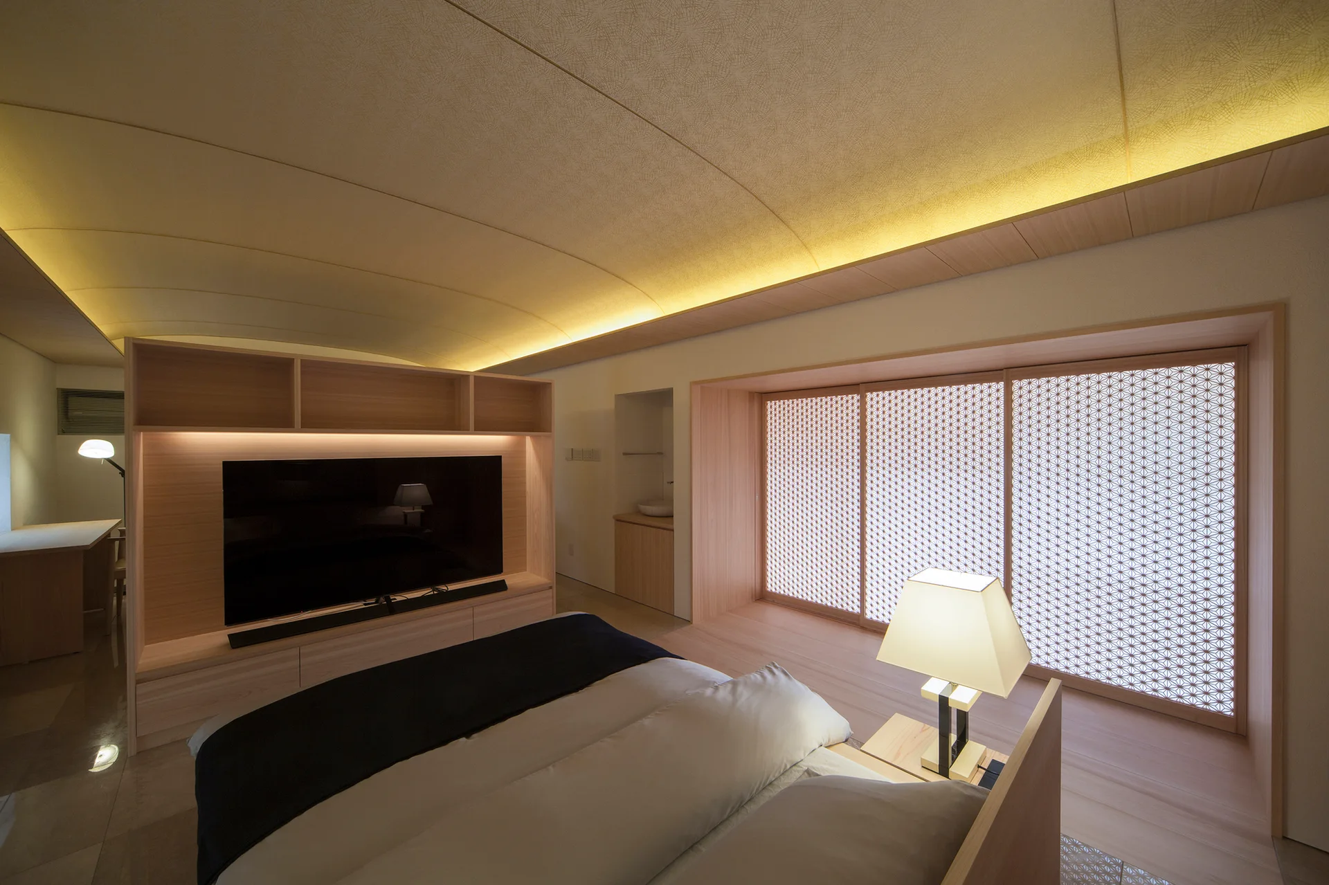 A minimalist bedroom features a low bed, a light wood media console, a curved ceiling with cove lighting, and patterned shoji screens.