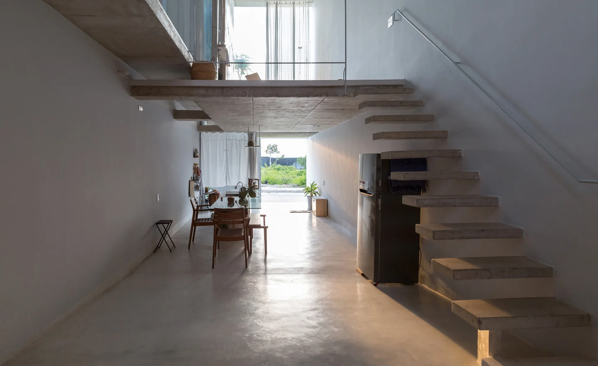 A minimalist home interior features a floating concrete staircase along a white wall, leading up to a concrete mezzanine overlooking a narrow dining area.