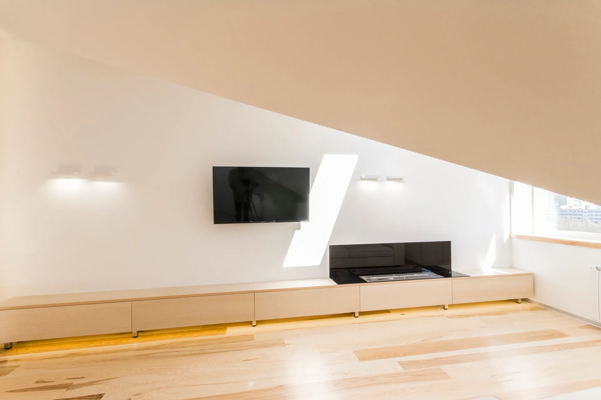 A minimalist media wall under an angled loft ceiling features a long light wood console, a mounted television, and a black glass fireplace insert.