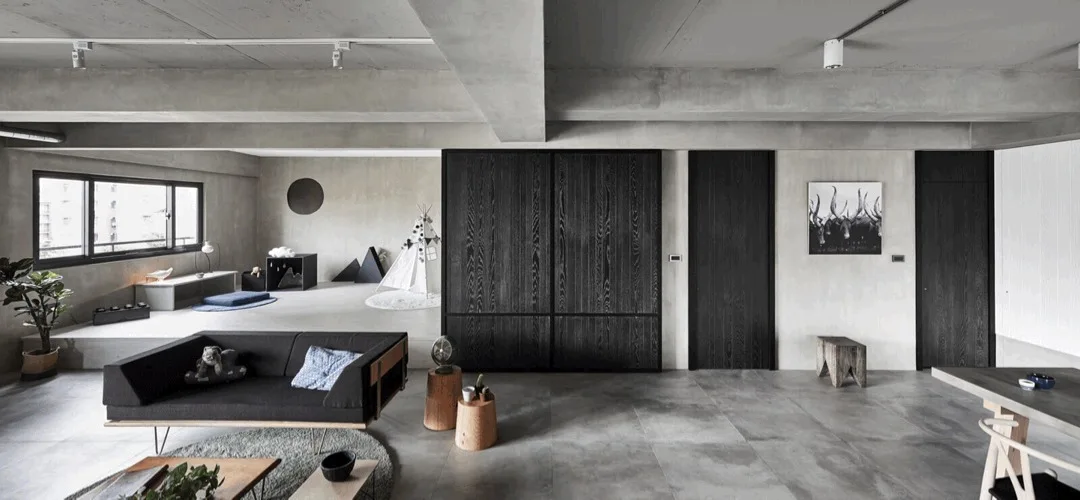 A minimalist concrete living room features a low-profile black sofa, dark wood cabinetry, and a raised platform play area with a teepee.