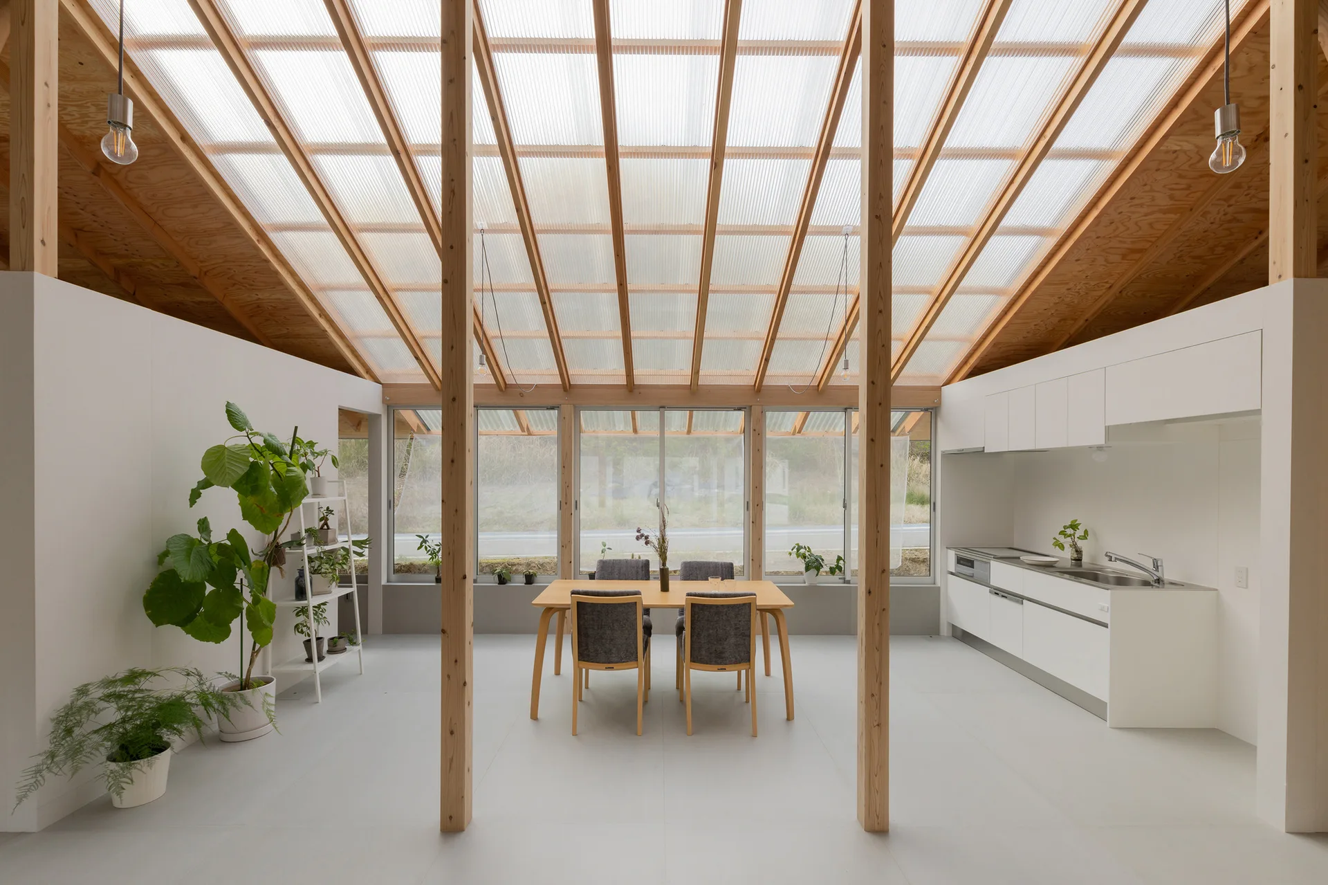 A minimalist dining and kitchen area featuring a sloped translucent roof with exposed timber columns, a wooden table, and white cabinetry.