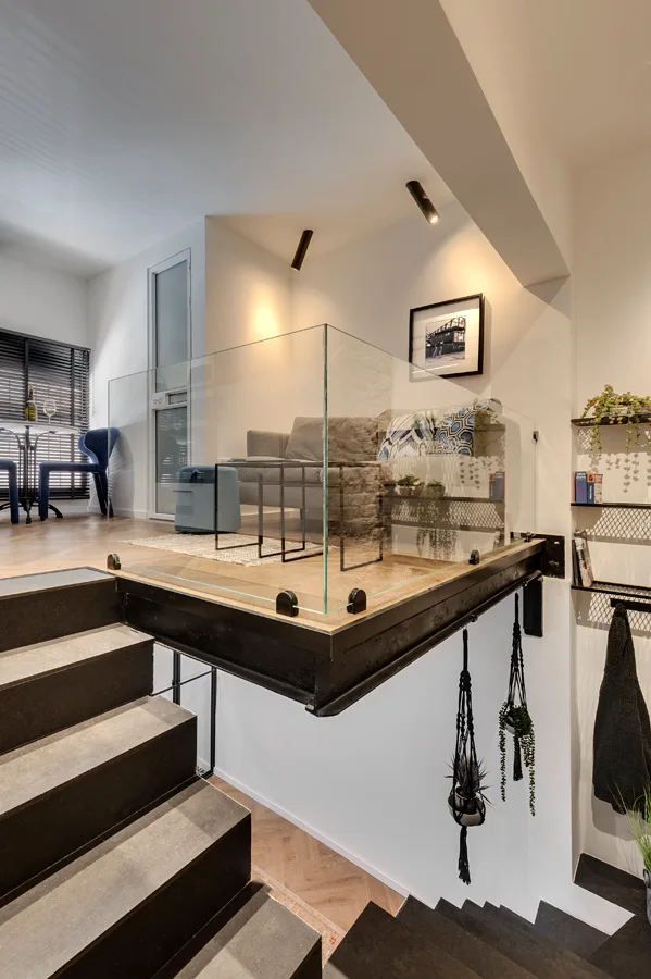 A dark staircase leads up to an elevated mezzanine living space with herringbone wood floors, a grey sofa, and a frameless glass railing.