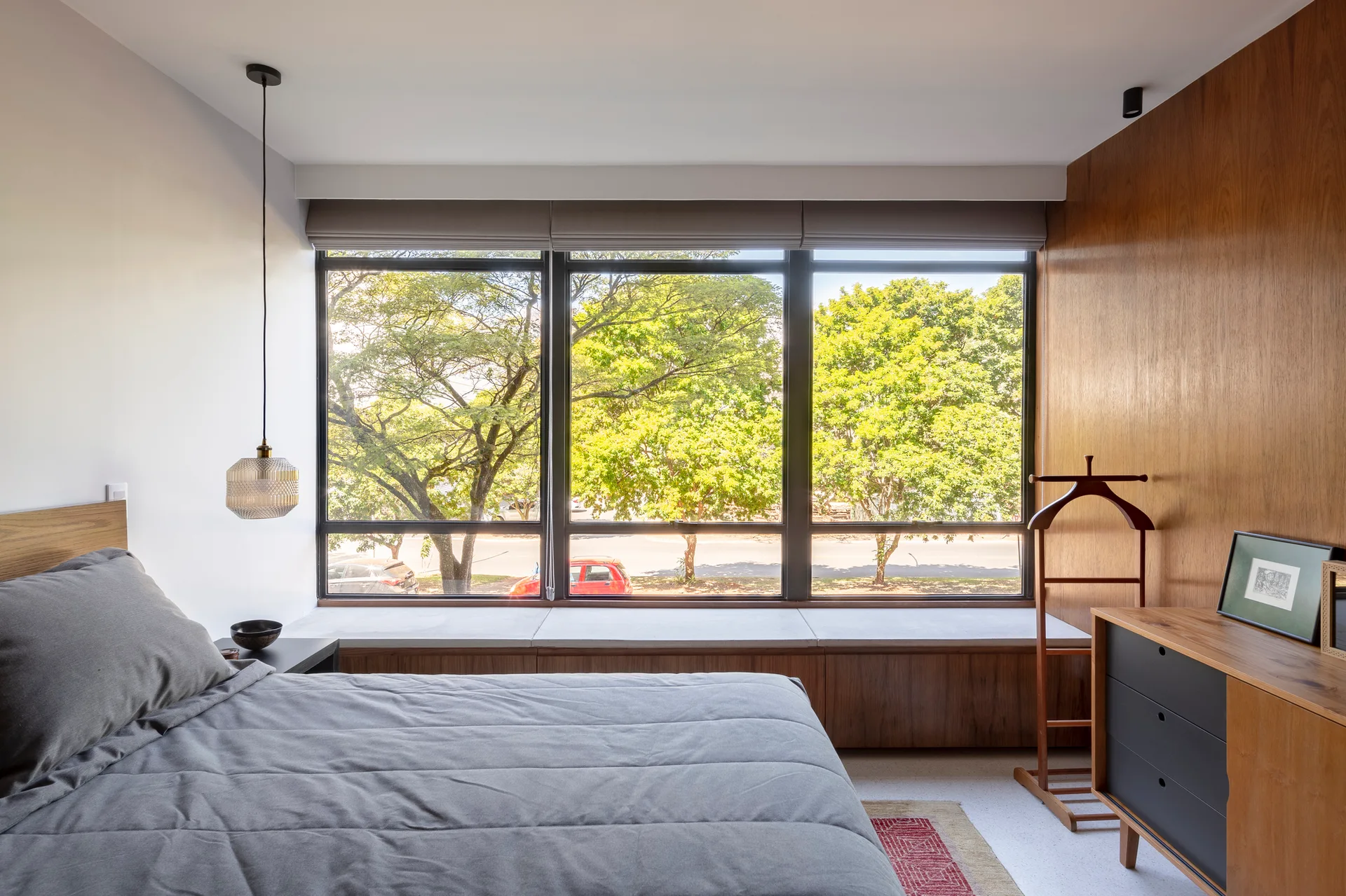 A modern bedroom with a grey bed, a long wood storage bench under a large window, and a warm wood paneled wall.