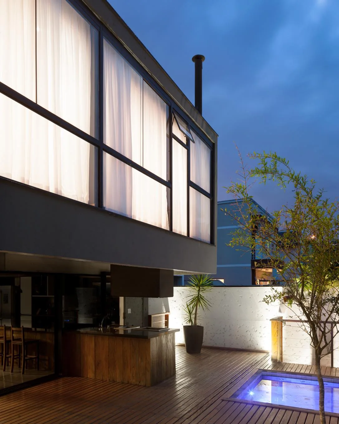 A modern backyard patio featuring a wooden deck, a small rectangular plunge pool, an outdoor kitchen, and large windows on the upper level at dusk.