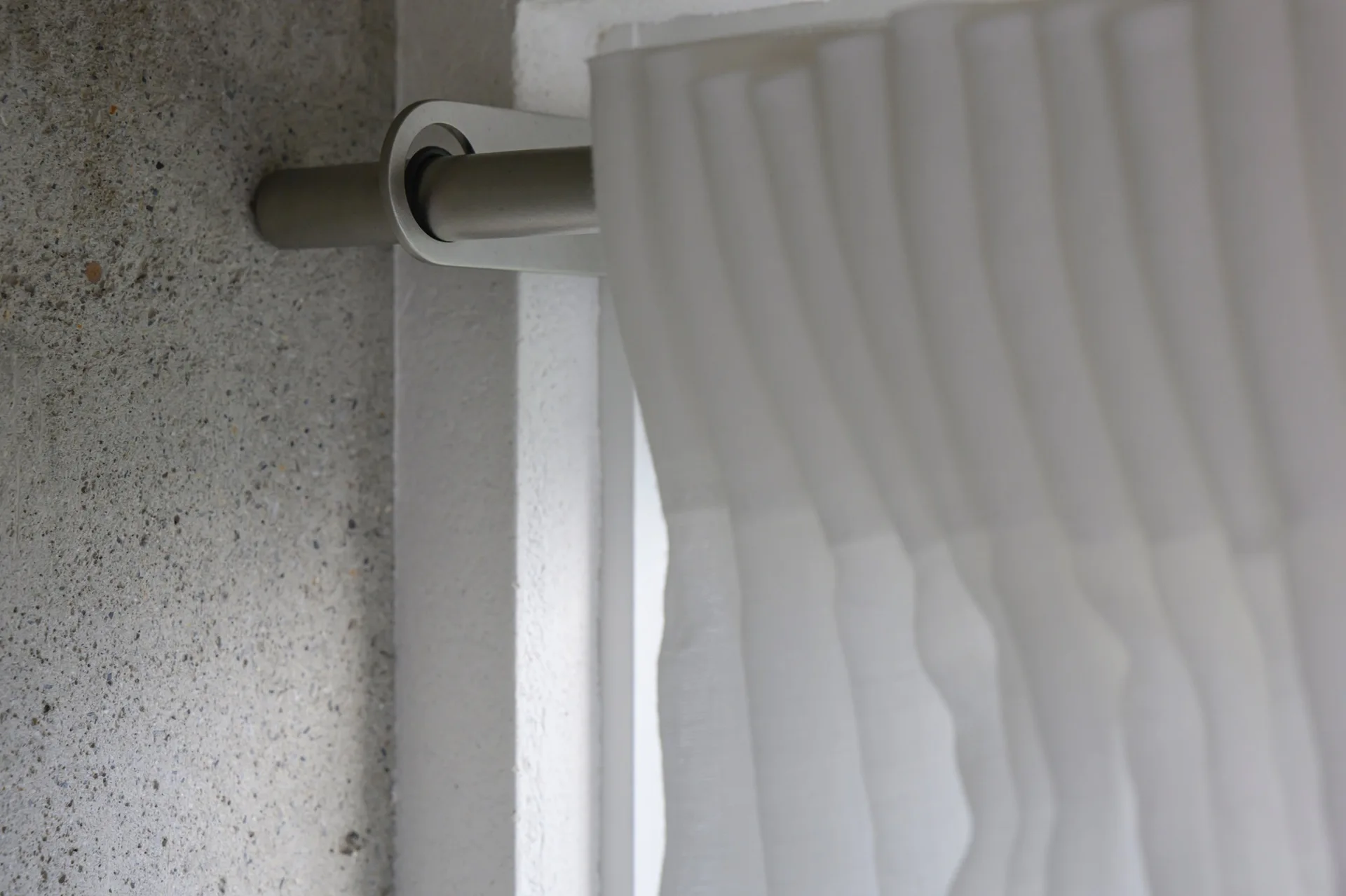 A close-up of a white pleated curtain hung on a matte metallic rod attached to a coarse textured grey concrete wall.
