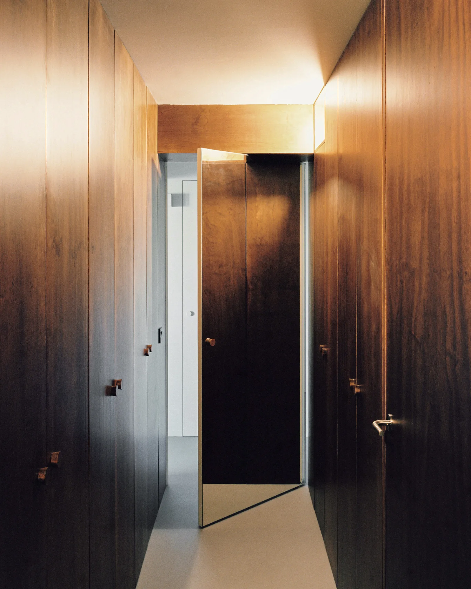 A narrow hallway featuring floor-to-ceiling dark wood wall paneling, a light grey floor, and an integrated wood pivot door swinging open.