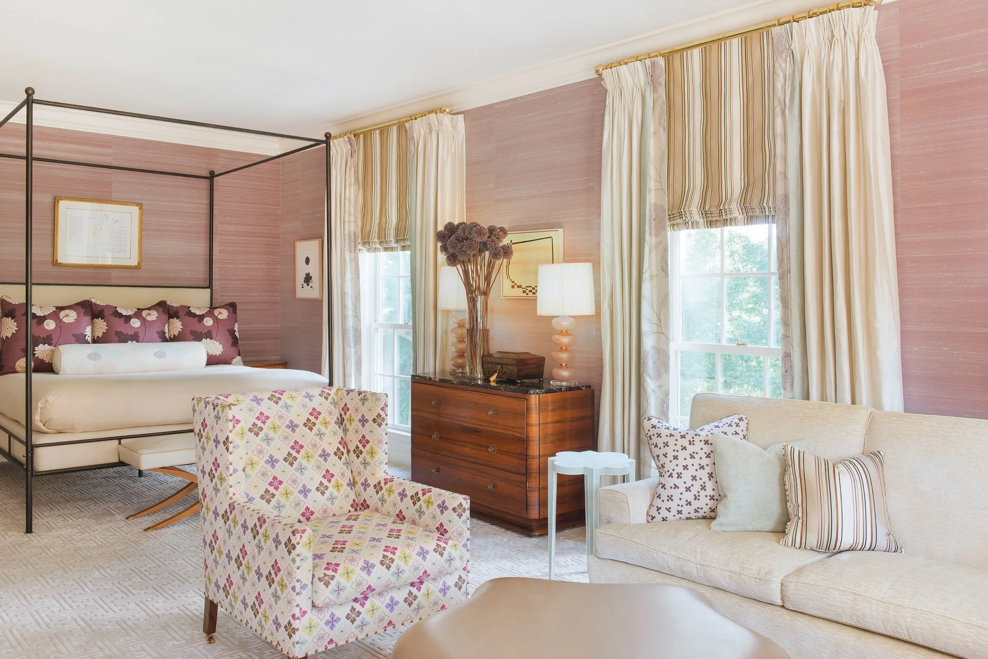 A spacious bedroom featuring a black canopy bed, a wooden dresser, and a sitting area with a floral armchair and a beige sofa.
