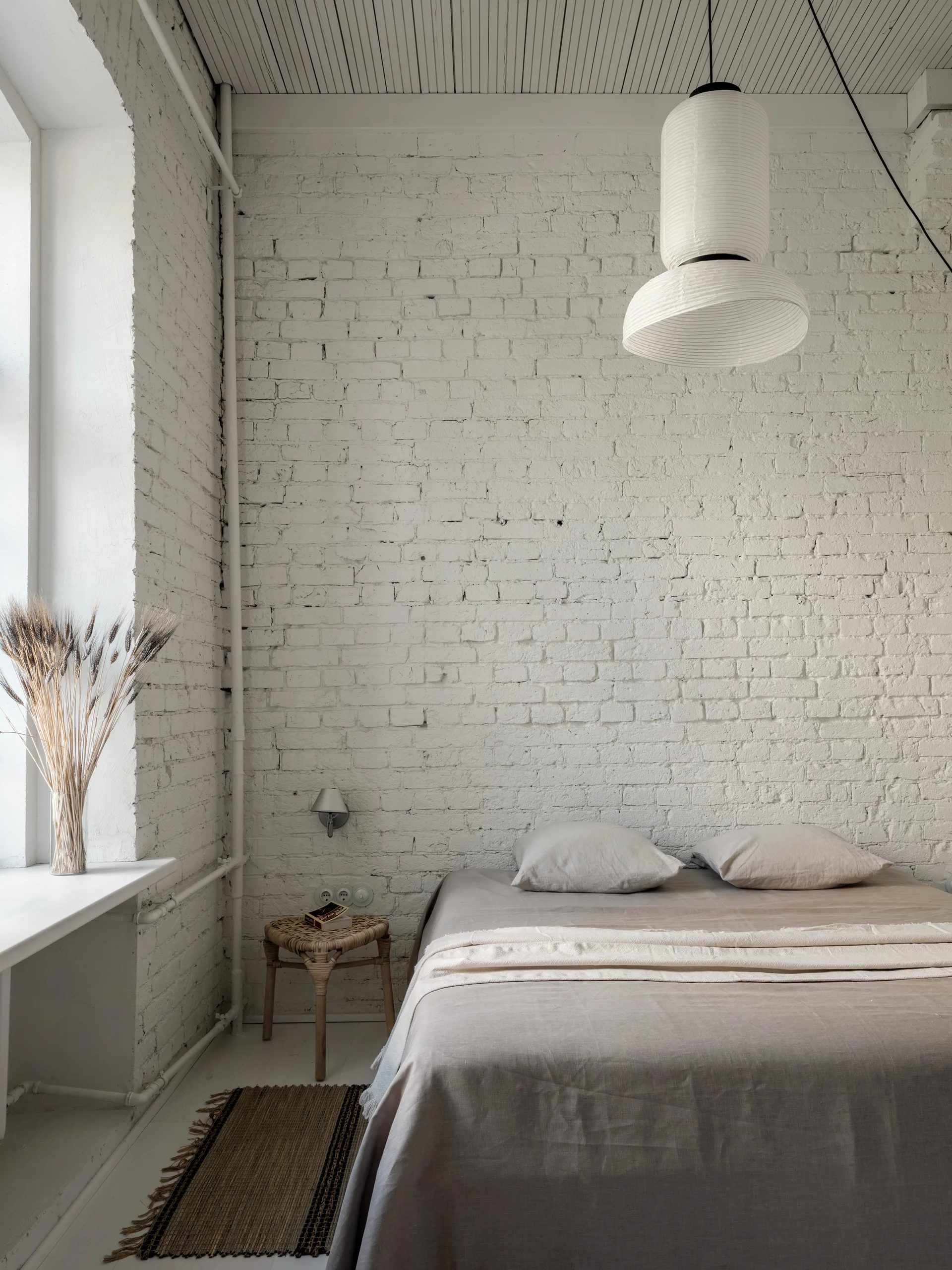 A minimalist bedroom featuring a bed with beige linen bedding against a painted white brick wall and under a large cylindrical paper pendant light.
