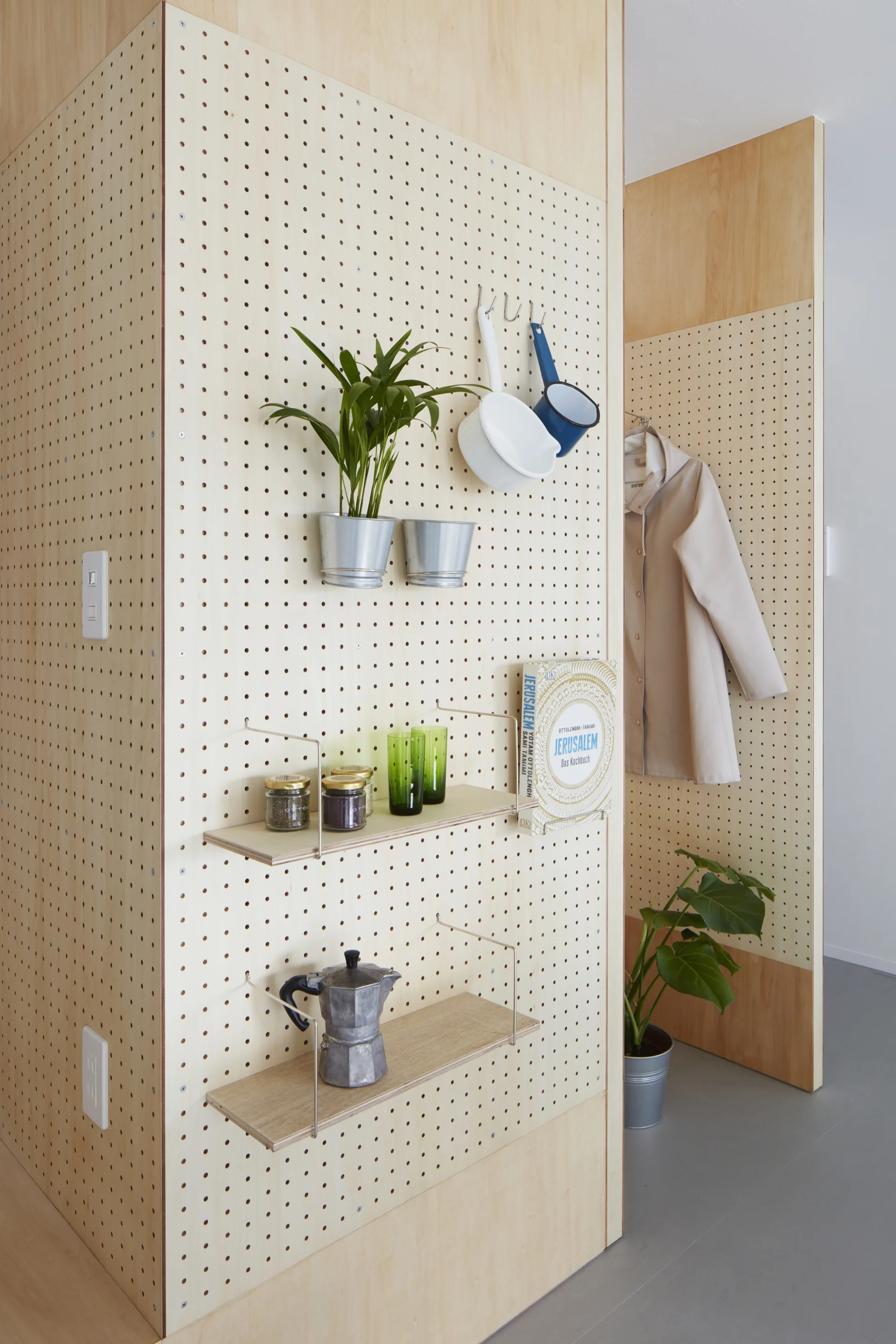 A light wood pegboard wall features thin adjustable shelves and hooks holding a moka pot, cookbook, potted plant, and hanging utensils.