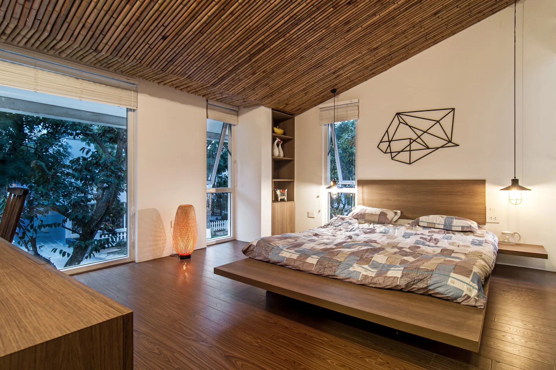 A modern bedroom features a low wooden platform bed, a sloped bamboo pole ceiling, and large windows looking out onto a garden.